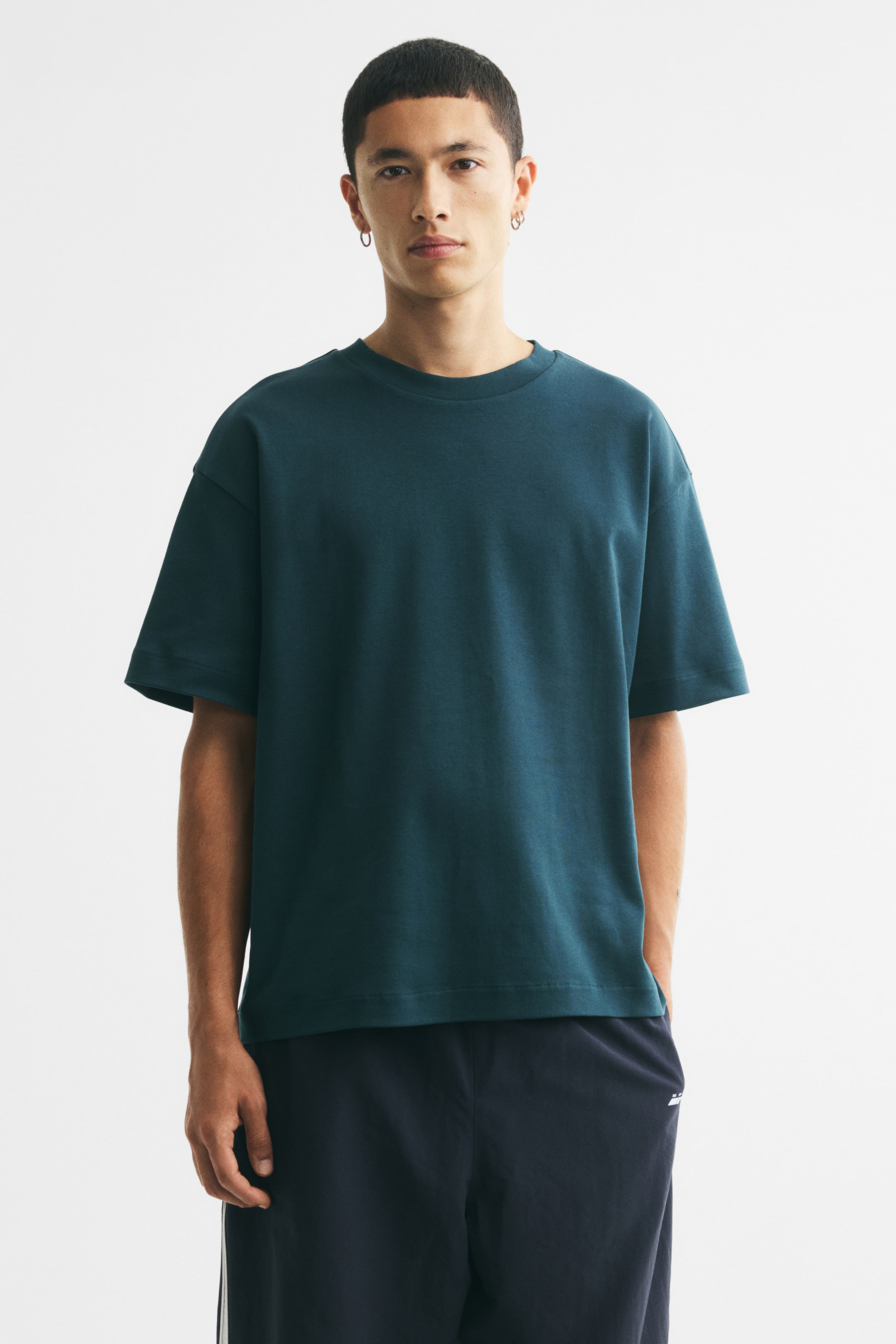 拡大画像を表示: コットンのインターロックジャージーを使用した、ダークティールのボクシーシルエットTシャツをモデルが着用しています。リブ仕上げのラウンドネック、ドロップショルダー、ストレートカットの裾が特徴で、ゆったりとしたフィット感を演出します。