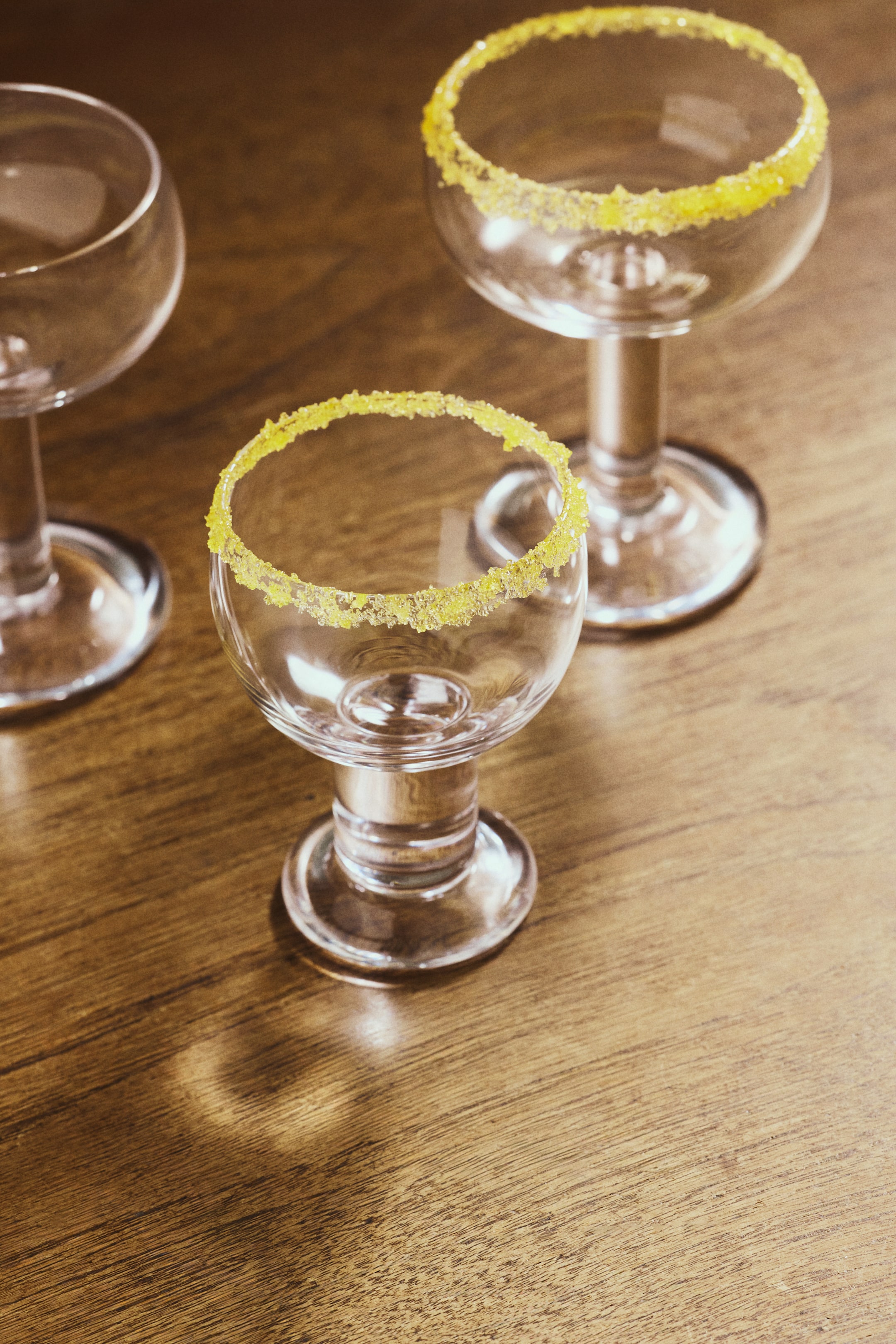 Voir une image plus grande: Trois verres à cocktail en verre transparent sont présentés d'une perspective légèrement surélevée, dont deux arborent un rebord de sucre jaune vif.