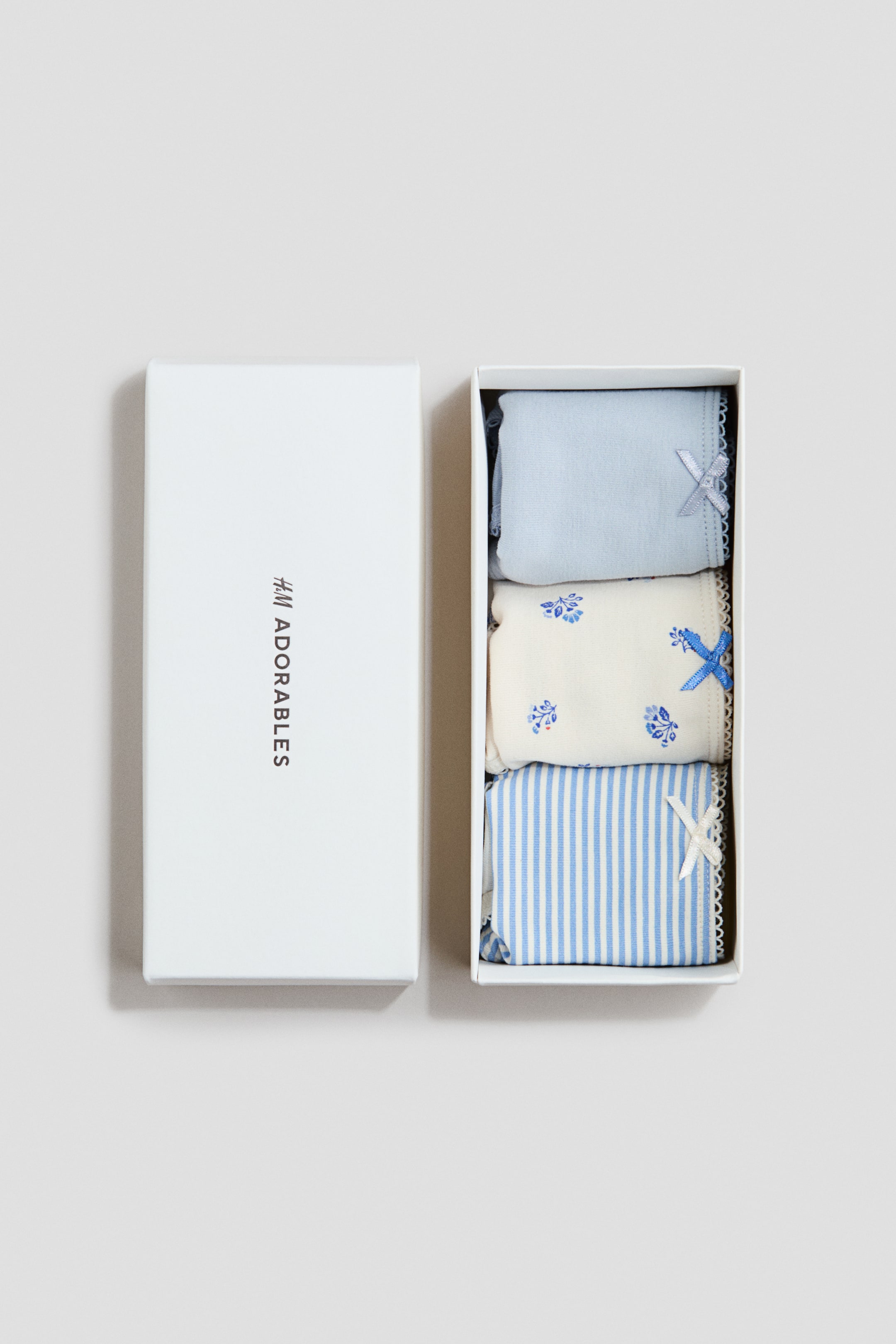 View larger image: Three H&M Adorables briefs are neatly arranged inside an open white box, showcasing their front designs. The top brief is a soothing light blue, adorned with a matching light blue satin bow at the front. The middle brief boasts an off-white hue with delicate blue floral patterns and a coordinating blue satin bow at the front. The bottom brief features a charming light blue and white striped pattern, accentuated by a pristine white satin bow at the front. Each brief is elegantly finished with white picot trims along its edges, adding a touch of sophistication.