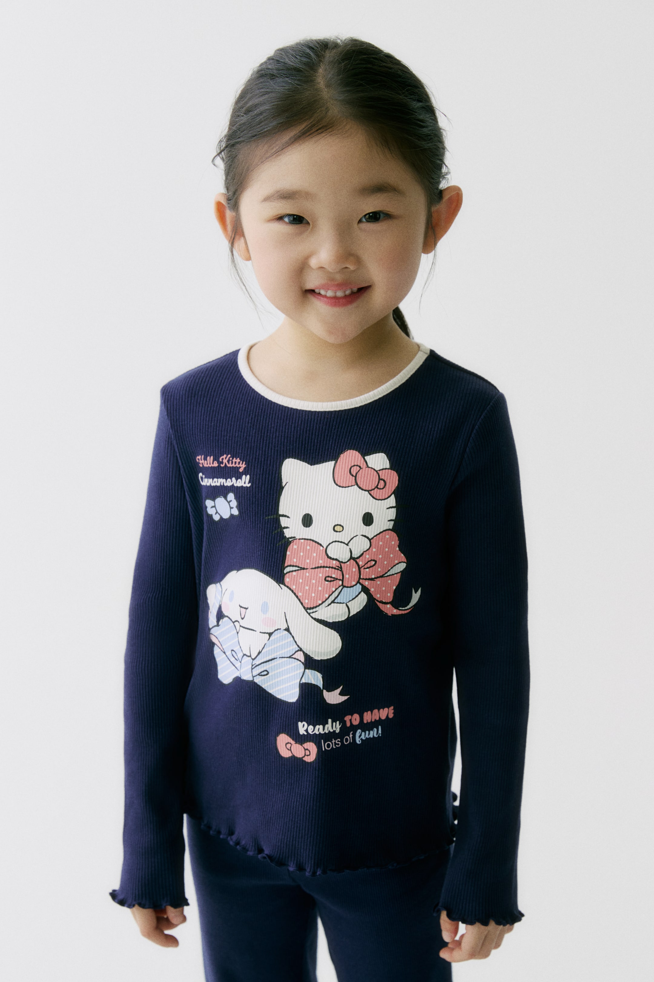 拡大画像を表示: ネイビーブルーのリブ編み長袖トップと同色のパンツを正面向きに配置しています。トップは白い縁取りのクルーネック、フリルの袖口と裾が特徴で、リボンを付けたHello KittyとCinnamorollのグラフィックプリントと、"Hello Kitty Cinnamoroll"および"Ready to have lots of fun!"のテキストが入っています。