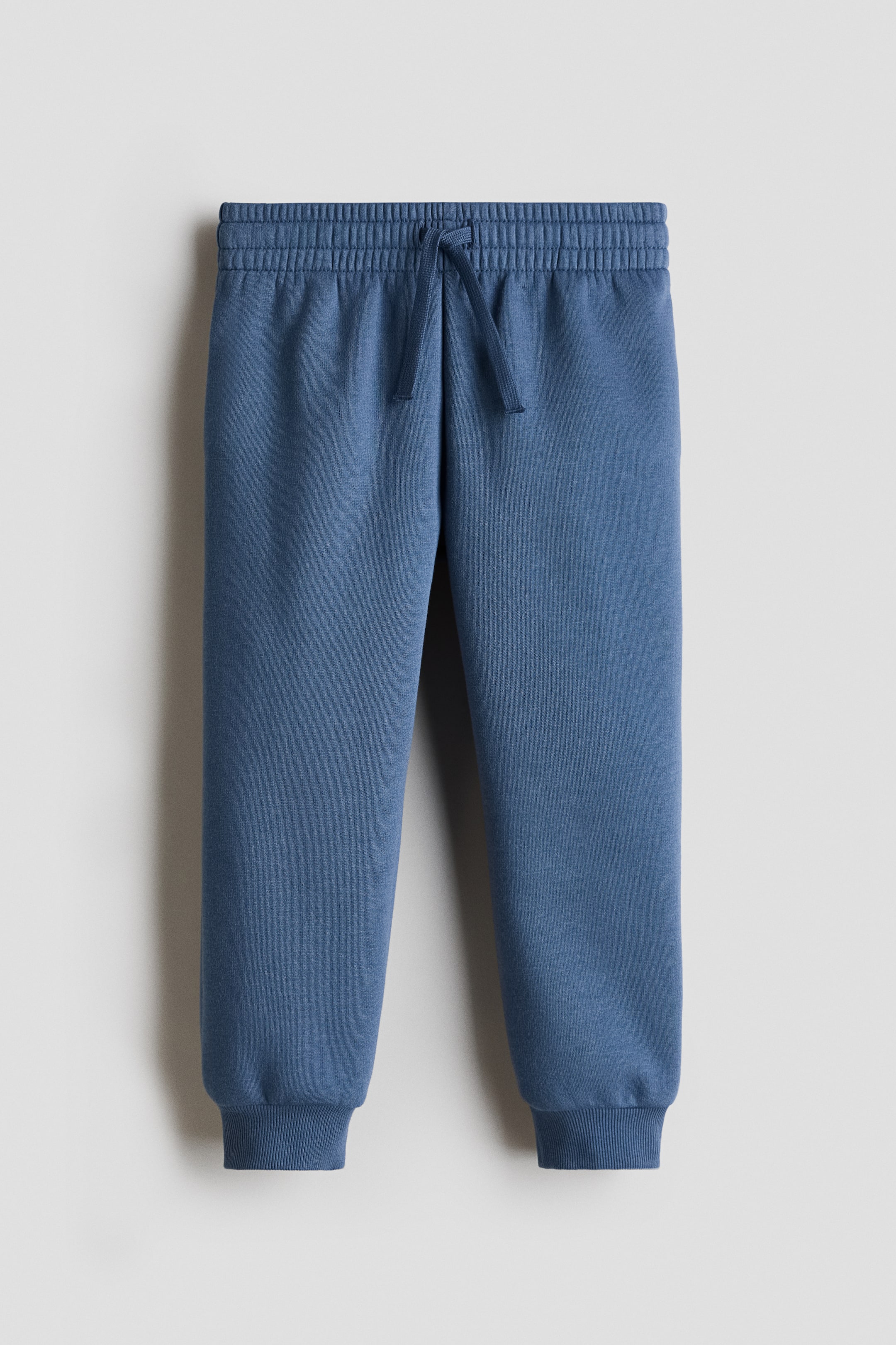 Vis større bilde: Blå joggebukser vises forfra, med en elastisk linning og en synlig snøring, og avsluttes med ribbestrikkede mansjetter ved anklene.