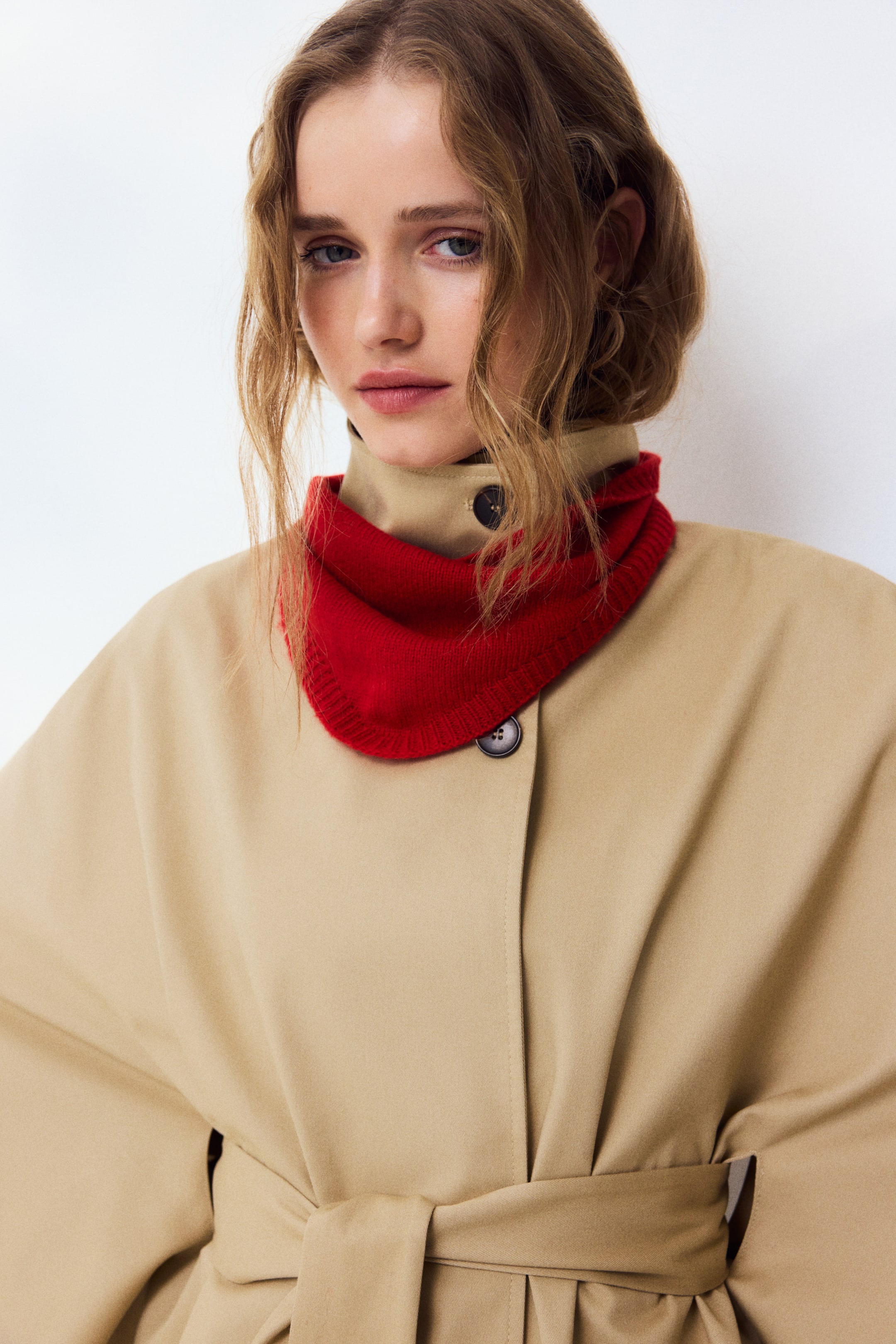 Grotere afbeelding bekijken: Een beige jas wordt gepresenteerd, met een opstaande kraag, een knoopsluiting aan de voorkant en een bijpassende zelfbindende riem die de taille accentueert. Een rode gebreide nekwarmer is gestyled rond de kraag van de jas.