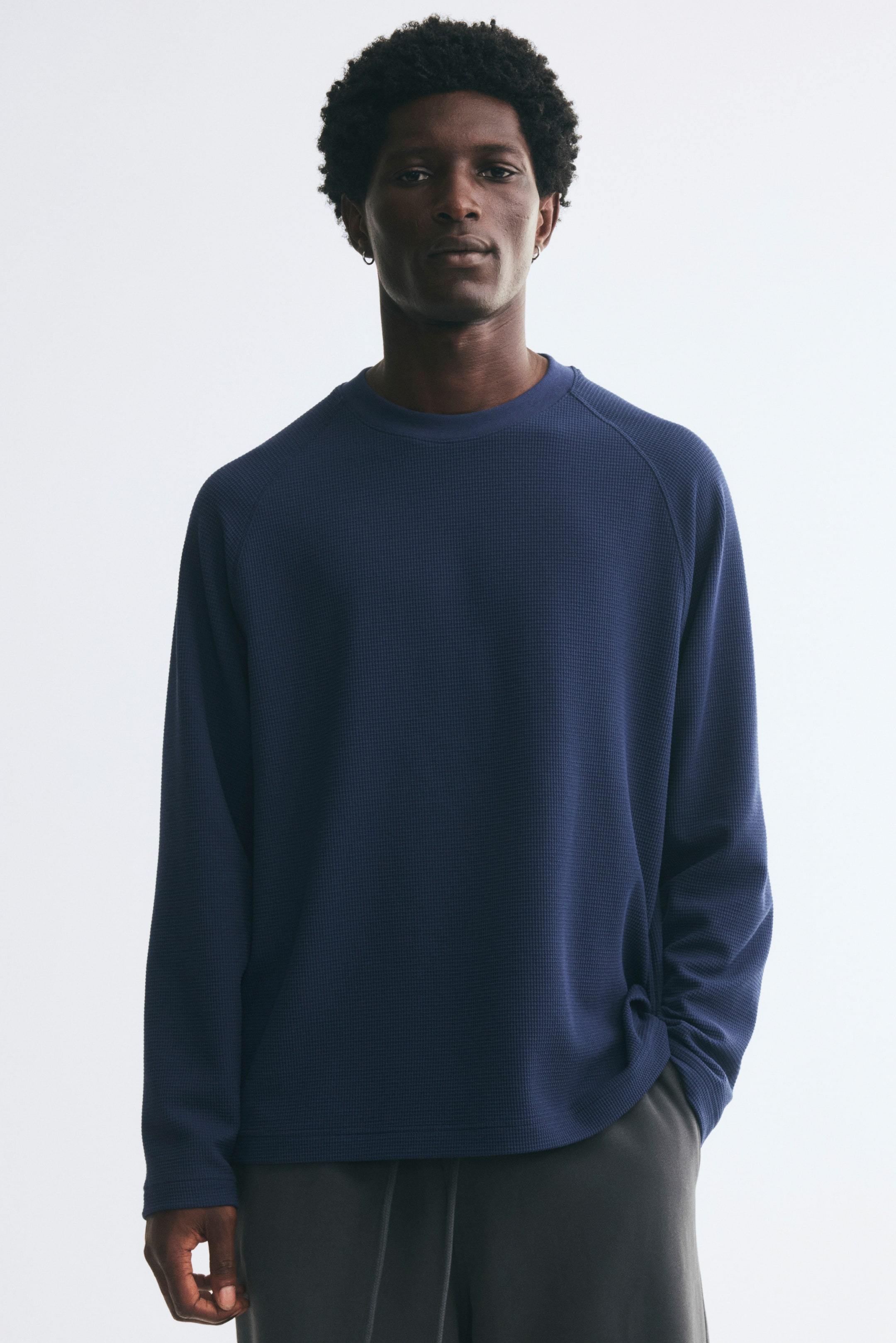 Voir une image plus grande: Un t-shirt en jersey gaufré bleu, à coupe décontractée, avec un col rond et des manches raglan longues, porté par le mannequin. Le t-shirt présente un ourlet droit et est agencé avec un pantalon gris foncé.