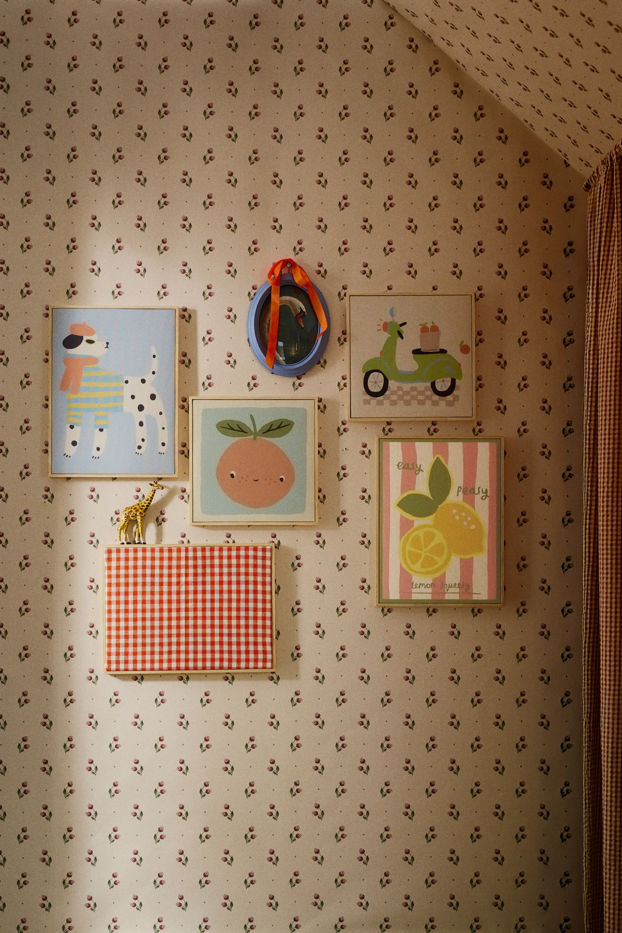 Büyük resmi görüntüle: Altı çerçeveli sanat eseri ve bir çerçeveli ayna, benekli bir köpek, gülümseyen bir turuncu meyve, yeşil bir scooter ve yazılı limonların renkli illüstrasyonlarını içeren bir galeri duvarı sergisi oluşturuyor. Kırmızı ve beyaz kareli bir çerçeve, desenini üst kenarında duran küçük altın bir zürafa figürü ile sergiliyor ve yuvarlak mavi bir ayna, beyaz bir kuğuyu yansıtarak ve turuncu bir kurdele ile süslenerek üst merkezde asılı duruyor.