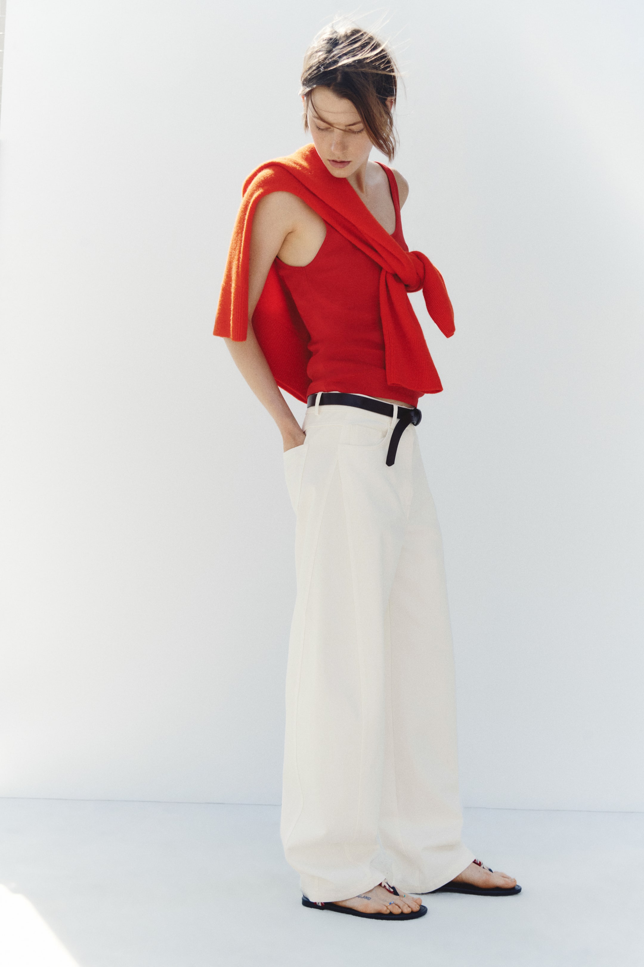 View larger image: Off-white, high-waisted denim jeans with curved, voluminous legs are shown facing left, highlighting their structured fit through the hips and thighs before tapering slightly at the hems. The jeans are styled with a thin black belt at the waist, a red sleeveless top, and flat sandals.