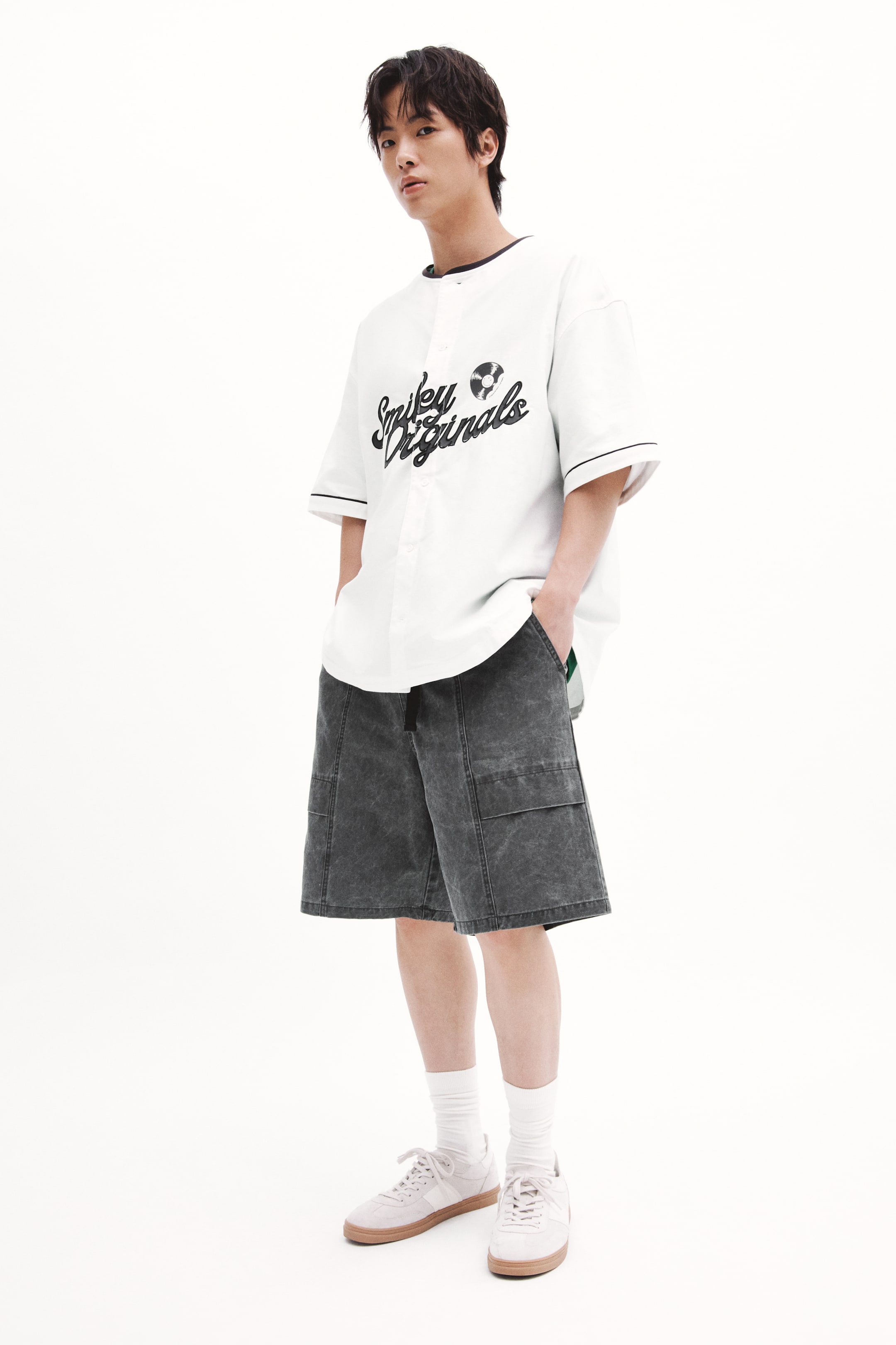View larger image: The model is donning a white short-sleeve baseball jersey featuring black trim and a 'Smiley Originals' graphic with a record icon. Paired with this is a set of dark grey acid-wash cargo shorts, equipped with side patch pockets, where the model's hands are casually nestled. The look is rounded off with light-colored sneakers adorned with white laces and complemented by white crew socks.