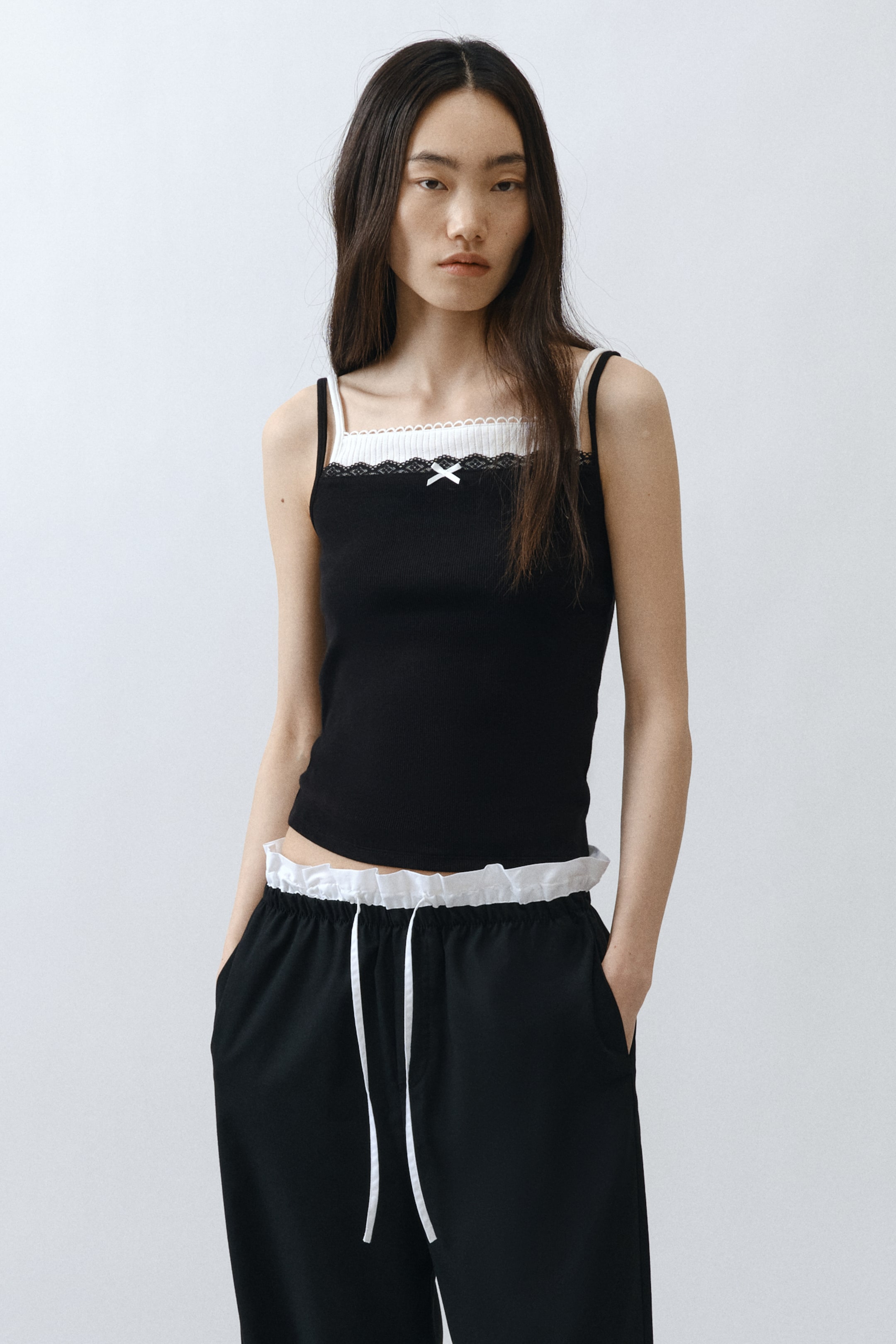 拡大画像を表示: モデルが着用しているのは、リブ編みのコットンジャージーで作られた、ショートでフィット感のあるストラップトップです。スクエアネックラインには黒のレーストリムが施され、フロントには小さな白いサテンのリボンがあしらわれています。さらに、白いポインテールジャージーのアンダーレイヤーにピコトリムが施され、追加のショルダーストラップが付いたレイヤードデザインが特徴です。