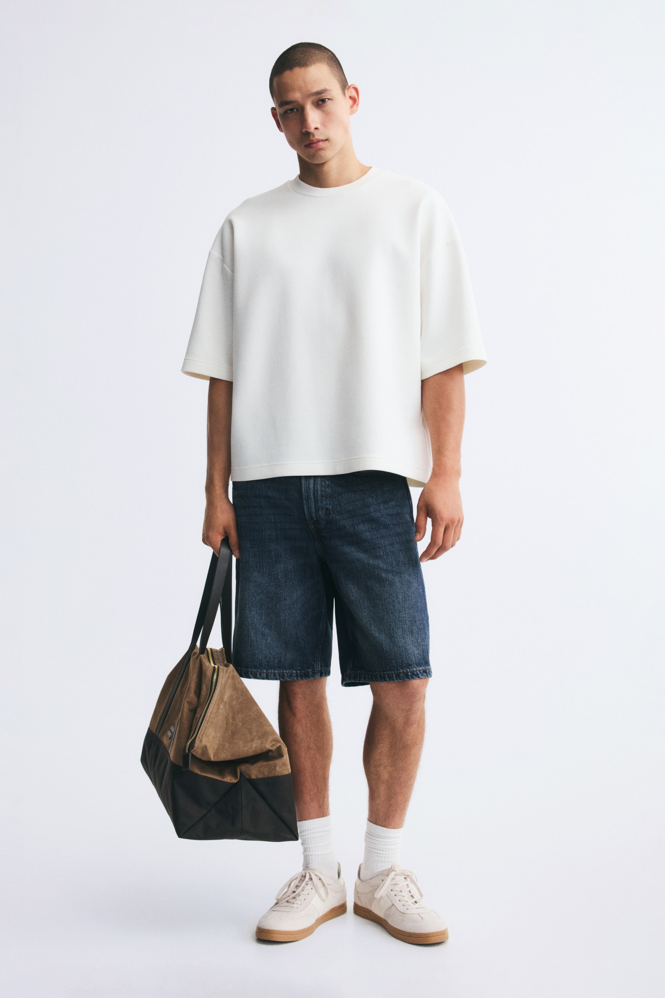 Vezi imaginea mai mare: Pantaloni scurți din denim albastru închis, cu croială lejeră și picioare conice care se opresc chiar sub genunchi, purtați de model. Pantalonii scurți sunt asortați cu un tricou alb supradimensionat, șosete albe, sneakers bej și o geantă maro și neagră ținută într-o mână.
