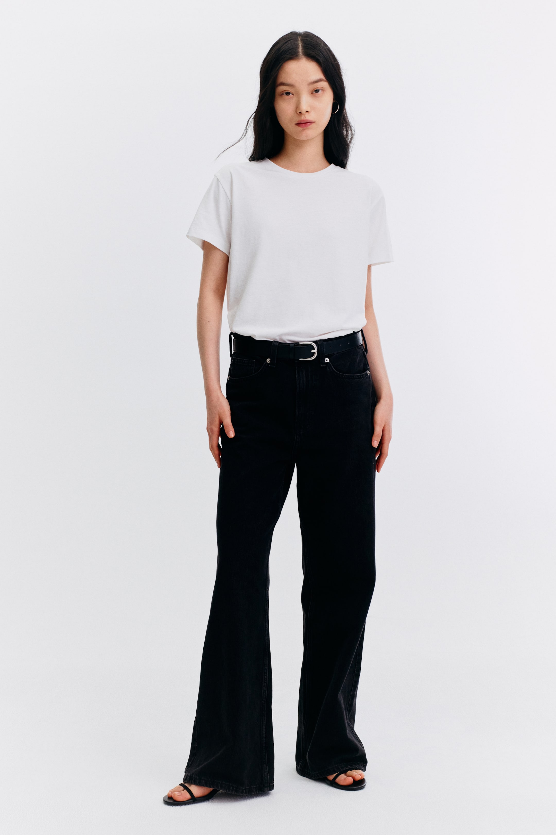 View larger image: The model is showcasing black flared jeans, complemented by a black belt and a white t-shirt neatly tucked in. She completes the look with black flat sandals, whose straps elegantly peek out from beneath the flared hem of the jeans.