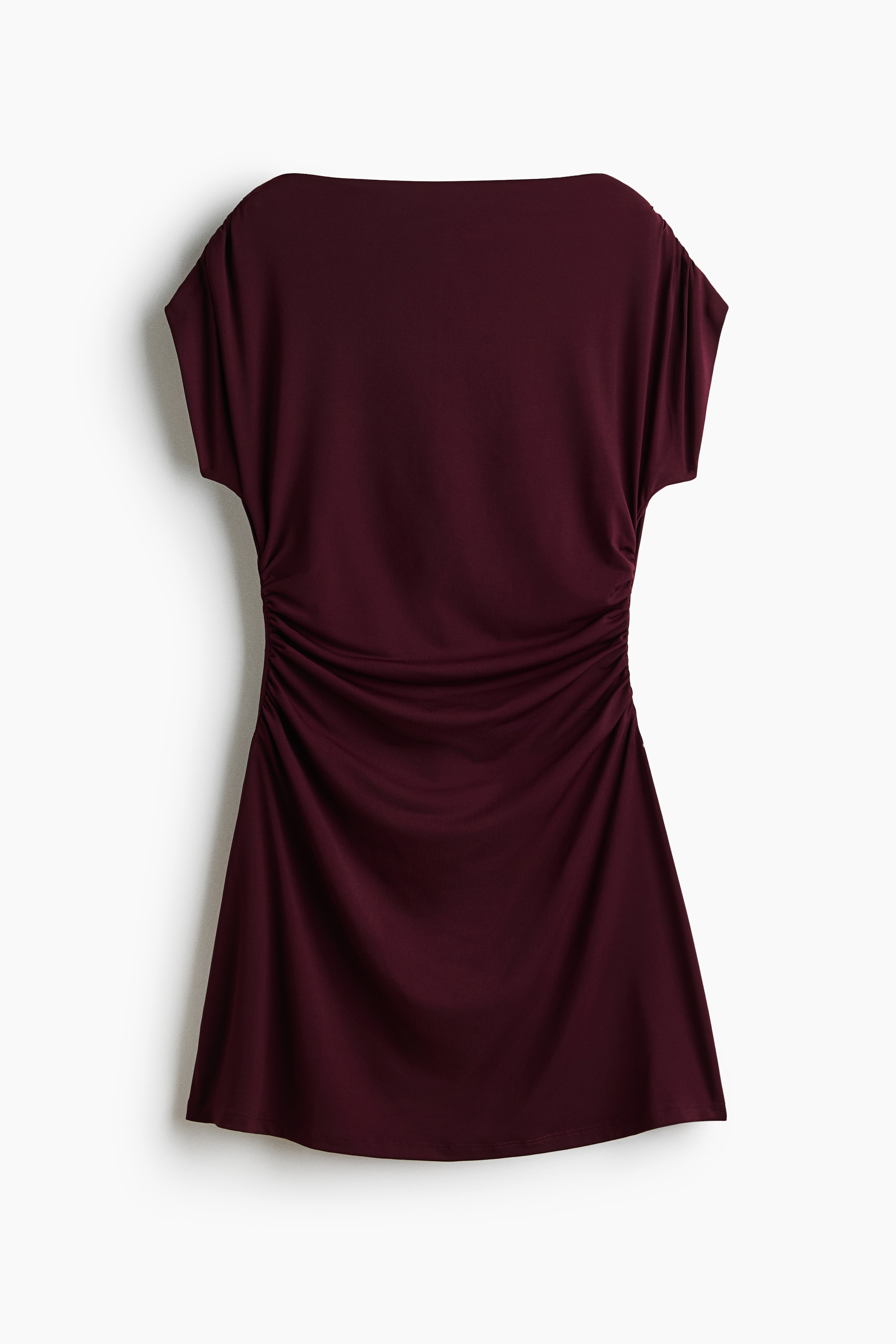 La modelo lleva un vestido corto de punto jersey color borgoÃ±a, con cuello barco, mangas cortas y costuras laterales fruncidas que crean un efecto drapeado. El vestido se combina con botas negras hasta la rodilla.