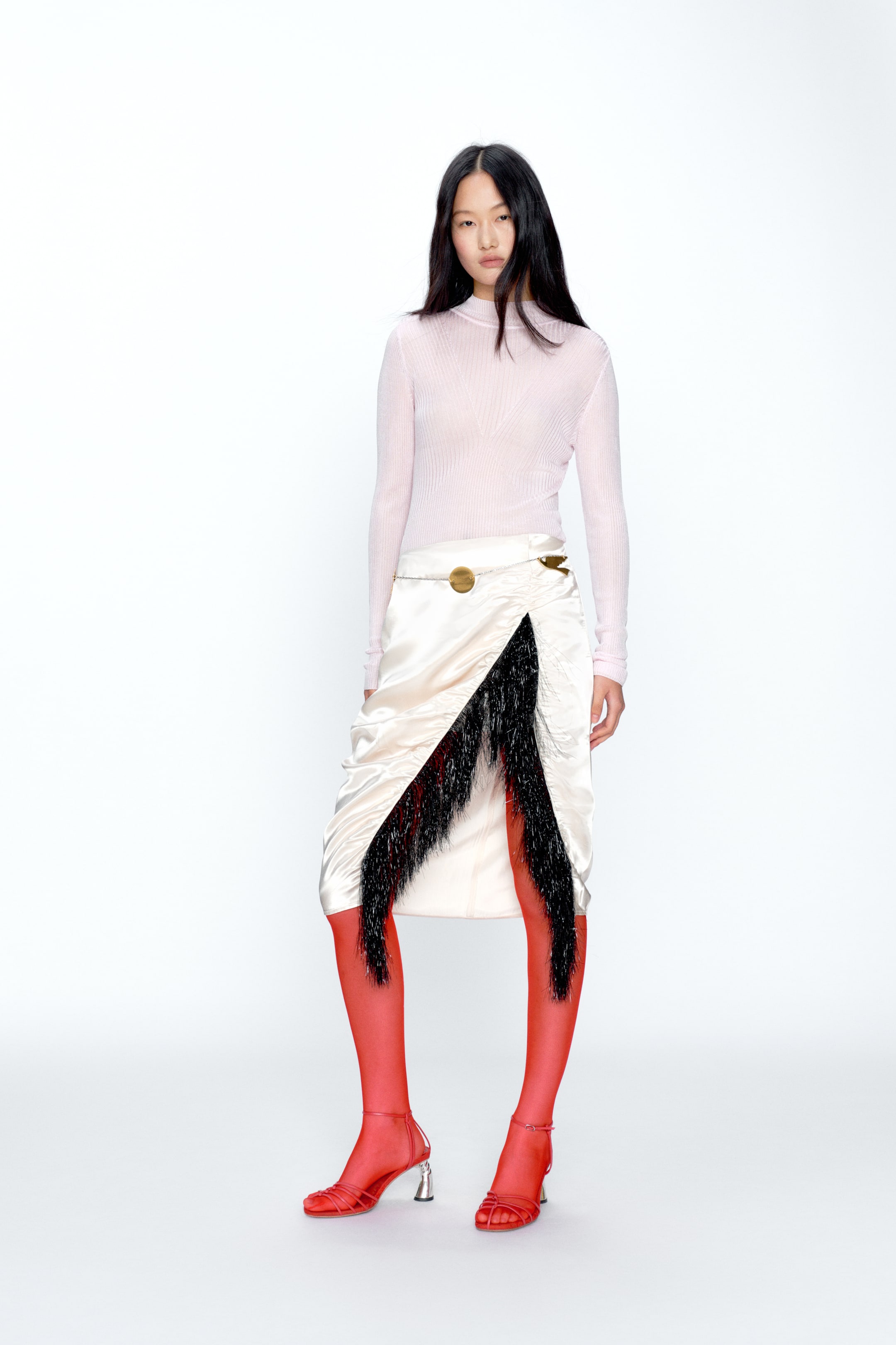 View larger image: A cream satin midi skirt featuring a draped, asymmetrical silhouette with a V-shaped front slit trimmed with black fringe. A slim gold-tone chain belt with a circular buckle accentuates the waist.