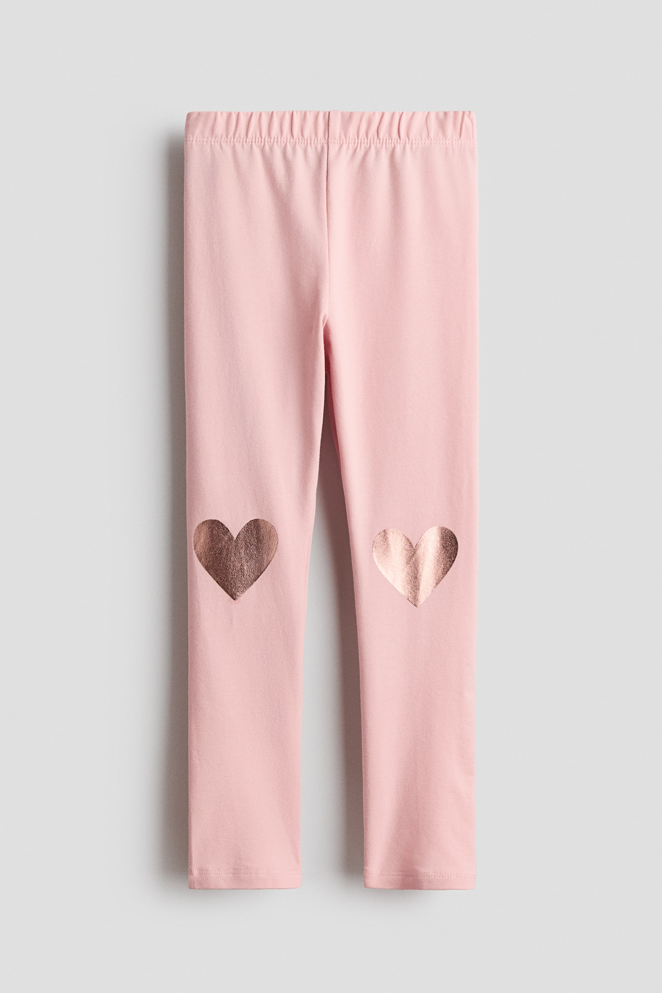 View larger image: "Front-facing pink leggings crafted from soft cotton jersey, featuring a covered elasticated waist and two metallic copper-coloured heart prints on the knee areas."
