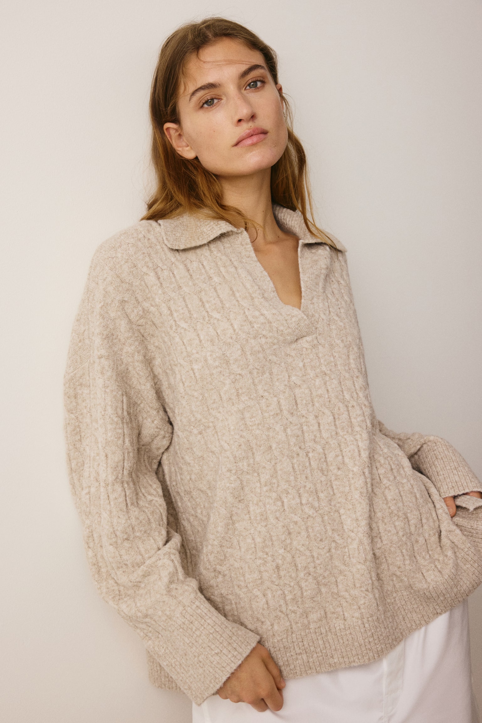 A beige cable-knit sweater is shown front-on, featuring a relaxed fit with a spread polo collar and an open V-neckline. The sweater showcases an all-over cable-knit texture, dropped shoulders, and ribbed trims, and is styled over white trousers. - 1