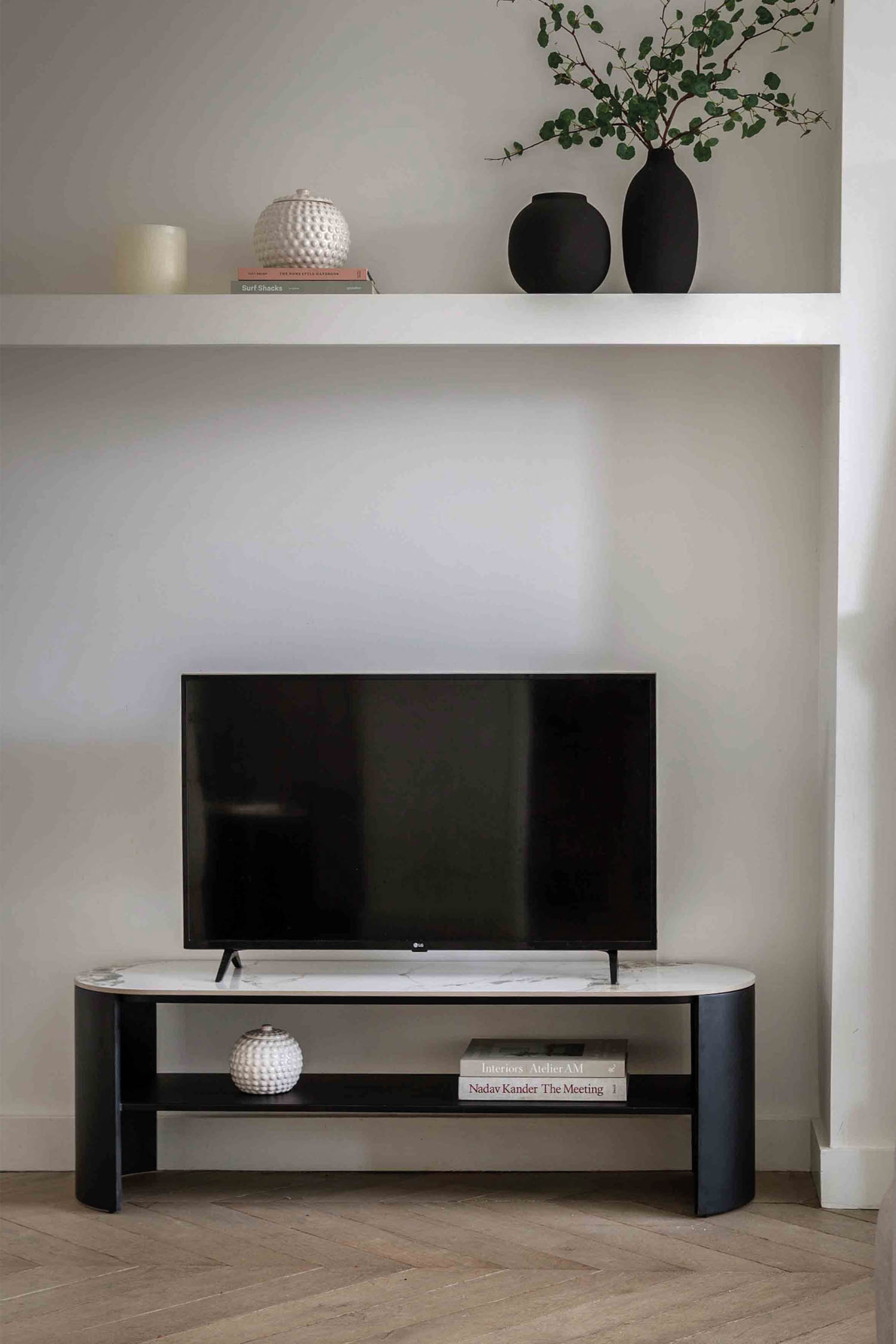 View larger image: A TV stand featuring a white marble-patterned oval top surface, upon which a black television screen is resting, is supported by two black curved side panels. A black rectangular lower shelf is positioned between these panels, accommodating a white textured spherical object and a stack of books.