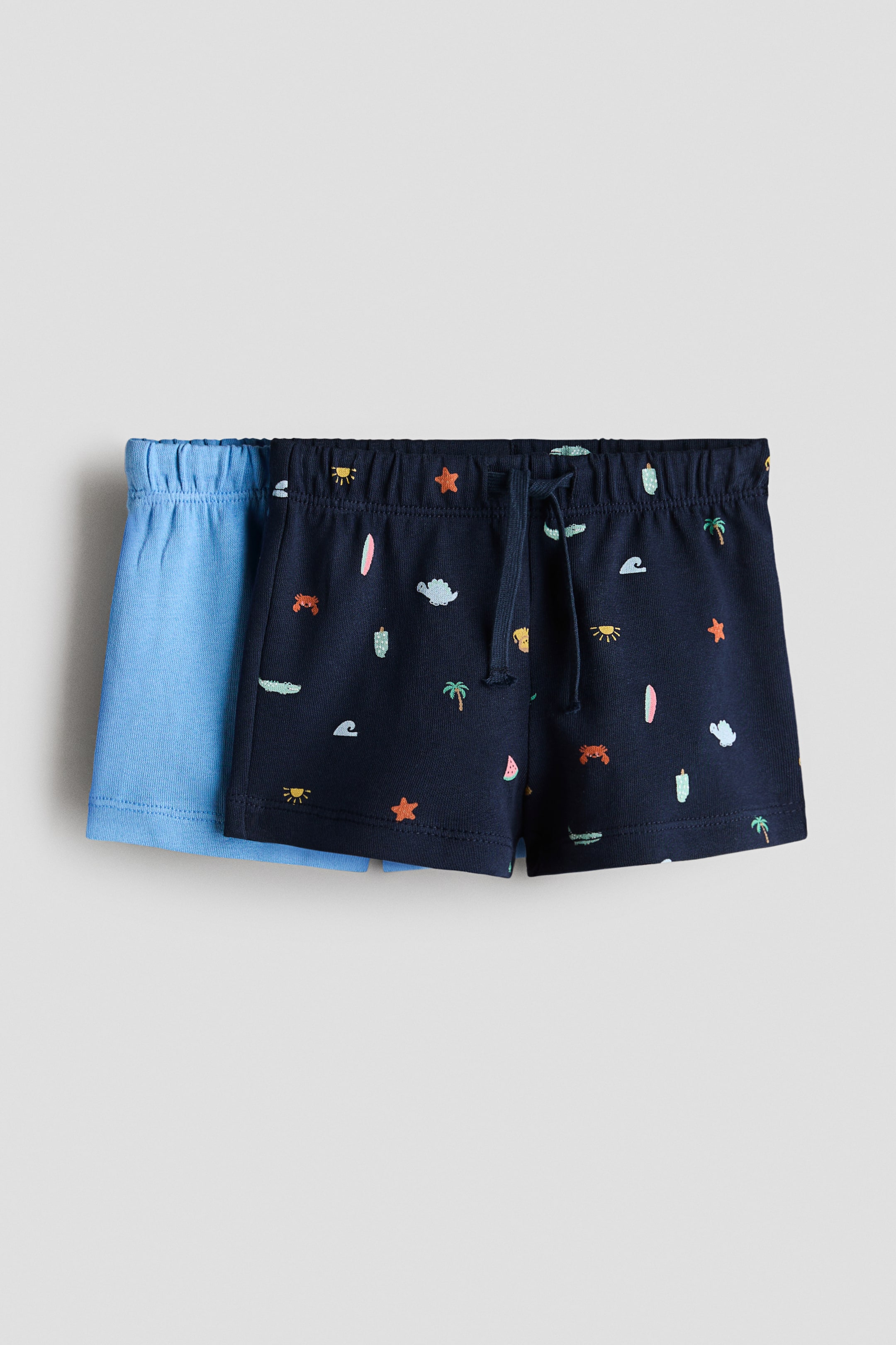 View larger image: "Soft, lightweight cotton sweatshirt shorts are showcased as a set of two, highlighting their front view. The navy blue pair, placed in the forefront, features a vibrant summer pattern and an elasticated drawstring waist with the drawstring neatly tied. Meanwhile, the light blue pair is partially positioned behind, also showcasing an elasticated waist."