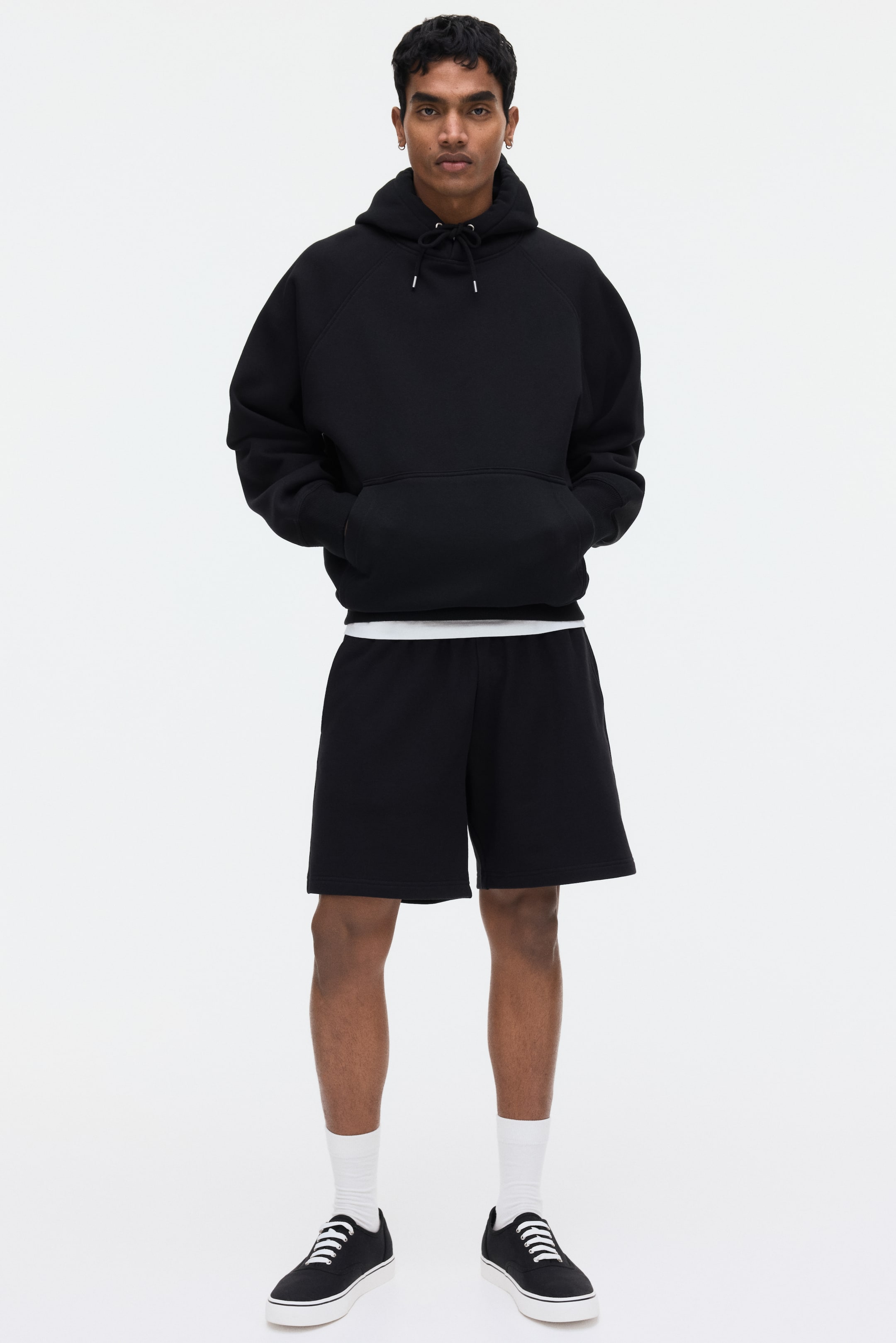 View larger image: The model is wearing black shorts made from a soft sweatshirt fabric. They have a regular fit, side pockets, and fall above the knees, paired with a white t-shirt visible at the waistline.
