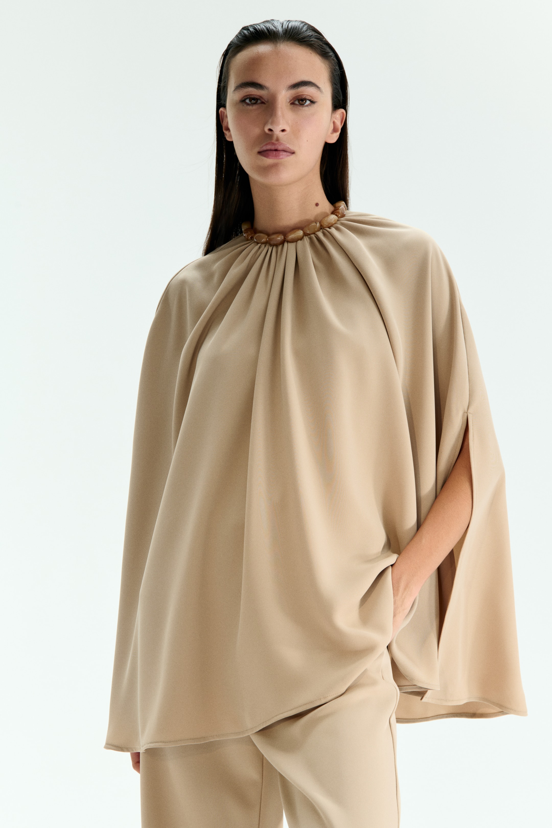 View larger image: A beige cape top is featuring a gathered high neckline with integrated light brown, rounded beads and loose sleeves with side slits. The model is wearing the top with matching beige pants, and one hand is resting in a pant pocket.