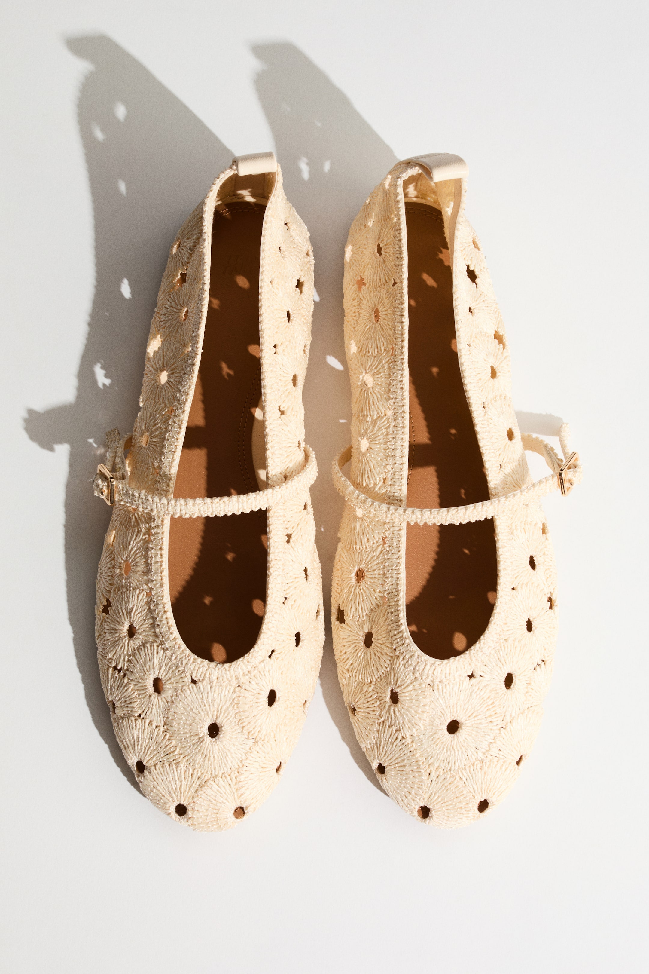 View larger image: A pair of light beige Mary Jane flats showcases a woven upper, intricately patterned with small, open-weave floral designs that create perforations. Each shoe features a thin strap across the instep with a small buckle and a slightly pointed toe.