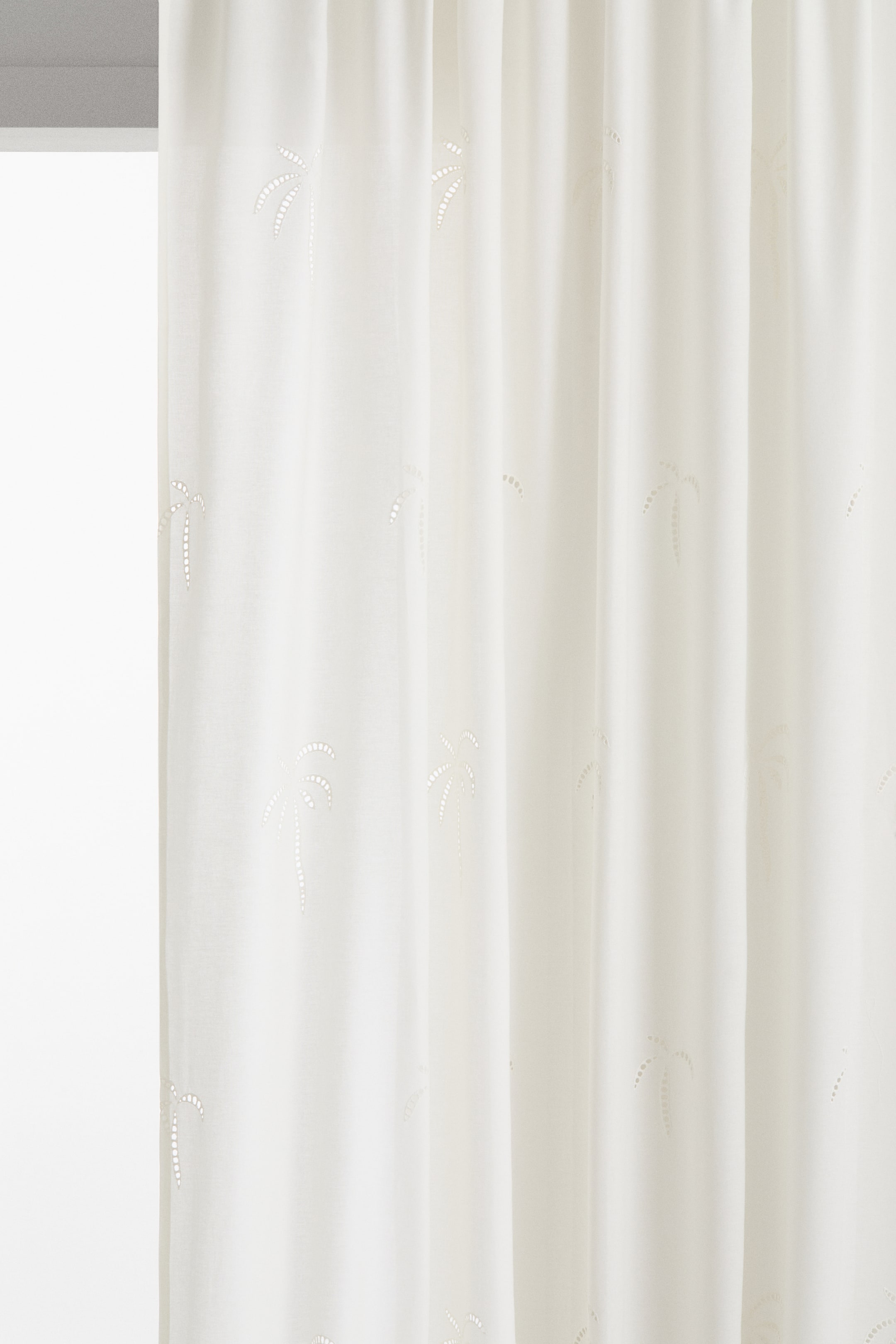 View larger image: A cream curtain showcases its front, adorned with a pattern of perforated palm tree shapes. The semi-sheer fabric drapes in gentle vertical folds.