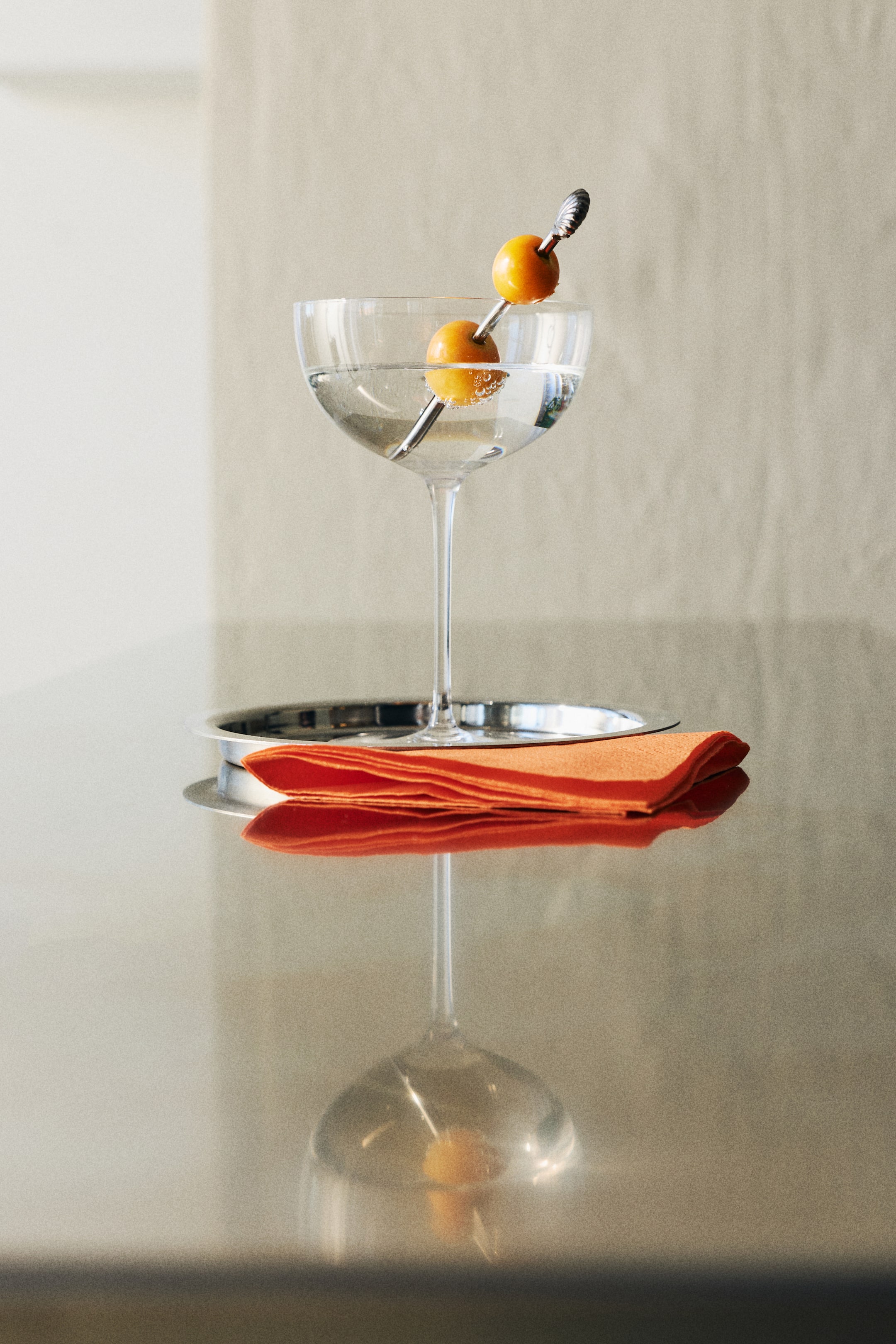View larger image: A clear coupe cocktail glass holds a translucent drink, adorned with a silver pick skewering two small orange spherical fruits. The glass rests on a silver round coaster, with a folded orange napkin placed beside it.