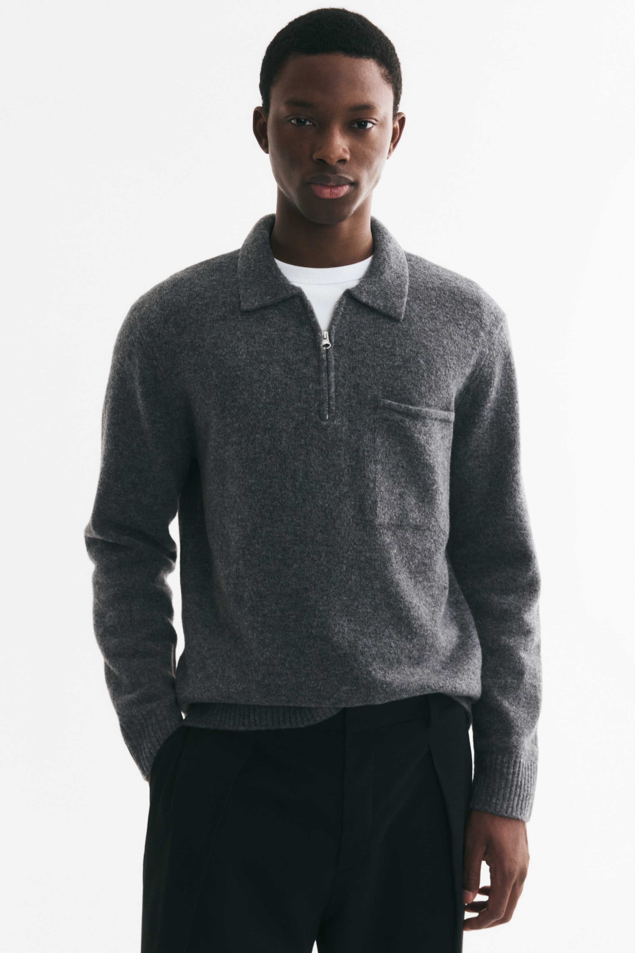 View larger image: A grey knit polo shirt featuring a collar, a half-zip neck closure, and a single chest pocket is worn by the model. The long-sleeved shirt is layered over a white T-shirt and features ribbed cuffs and a ribbed hem.