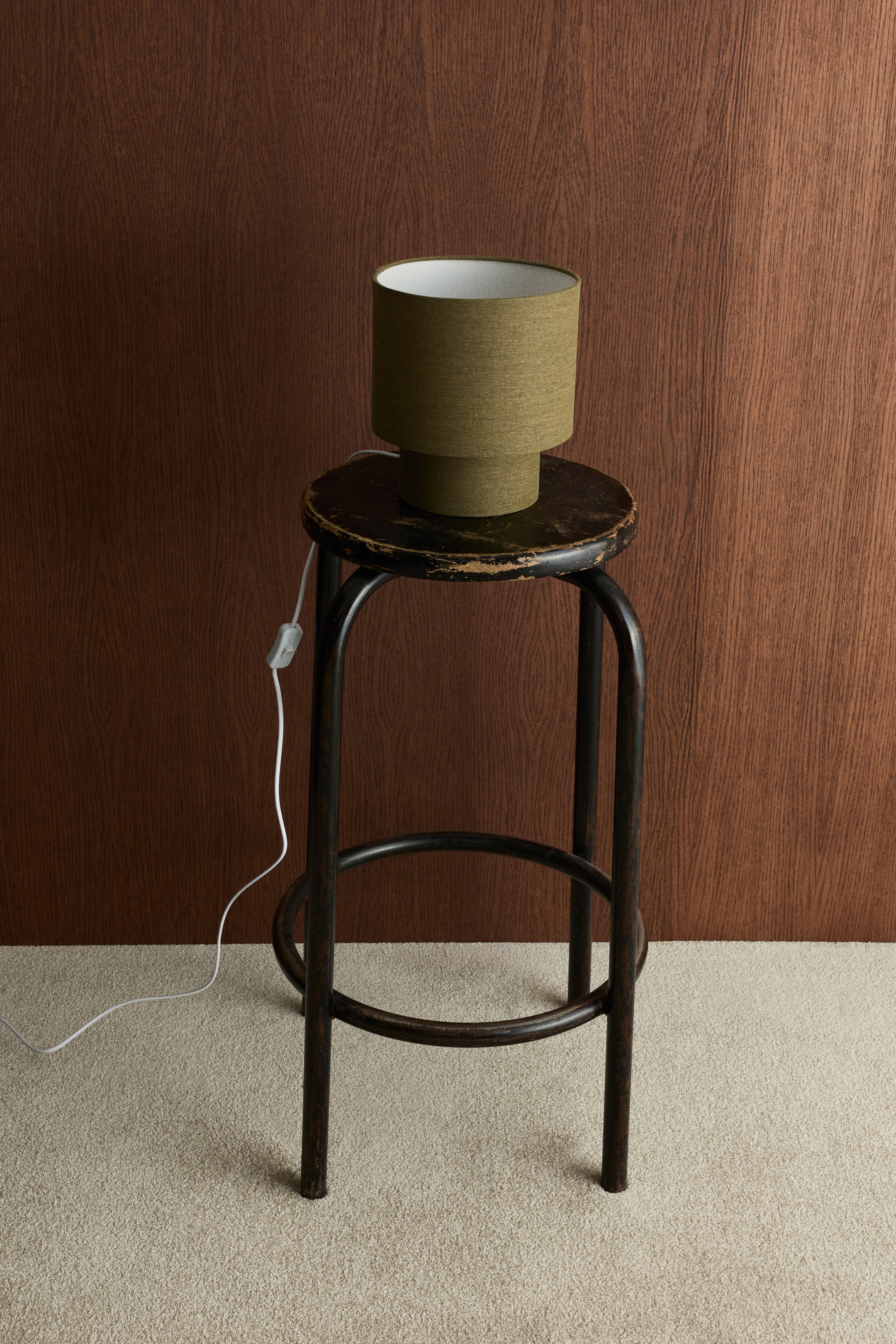 View larger image: A lamp featuring a cylindrical olive-green fabric lampshade with a white interior sits atop a slightly narrower, matching cylindrical base. A white mains cable with an inline switch extends from the lamp base.