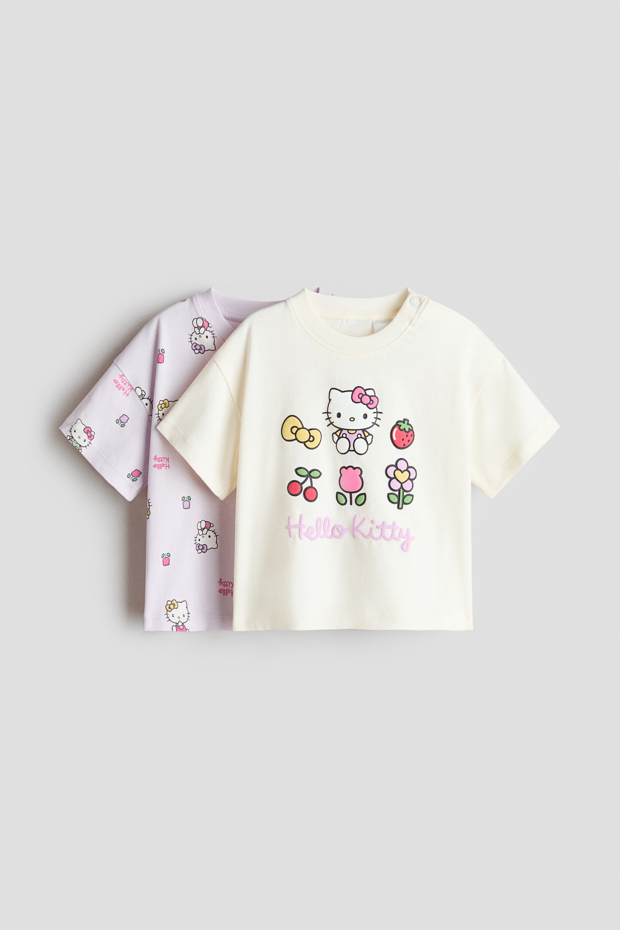 View larger image: Visible here is a 2-pack of short-sleeved cotton T-shirts, featuring a cream shirt with a ribbed neckline, adorned with a large front graphic print of Hello Kitty and other motifs, complemented by a snap button closure on its right shoulder. Additionally, the pack includes a lavender shirt with a ribbed neckline, showcasing an all-over pattern of smaller Hello Kitty designs.