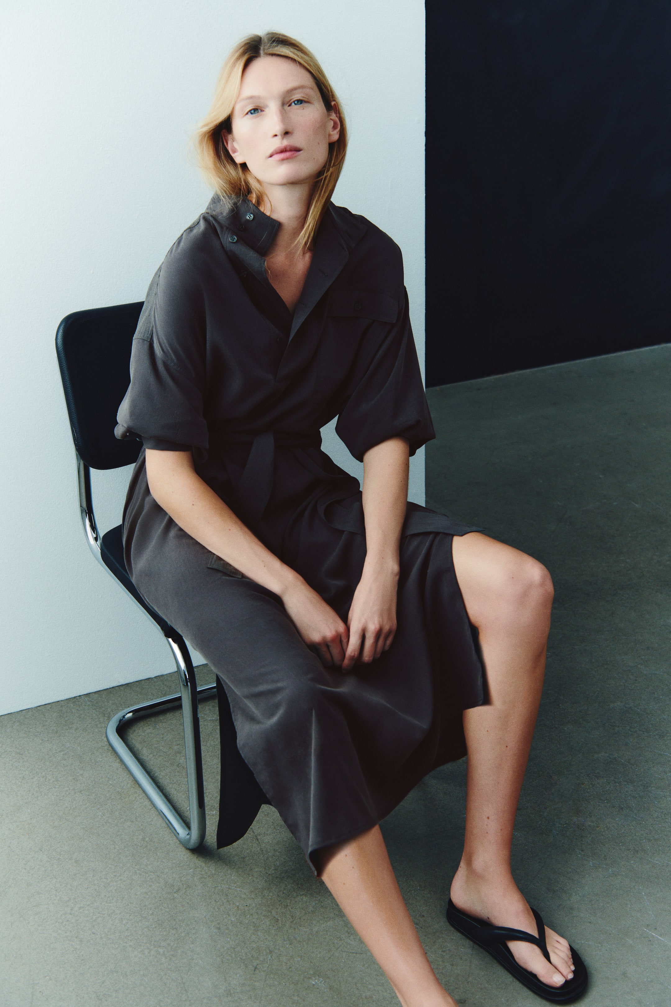 View larger image: A dark grey long-sleeve tie-belt dress is being worn by the model sitting on a chair, with the sleeves pushed up to the elbows. The midi-length dress features a high buttoned collar, a self-tie waist belt, and a side slit, and is styled with black thong sandals.