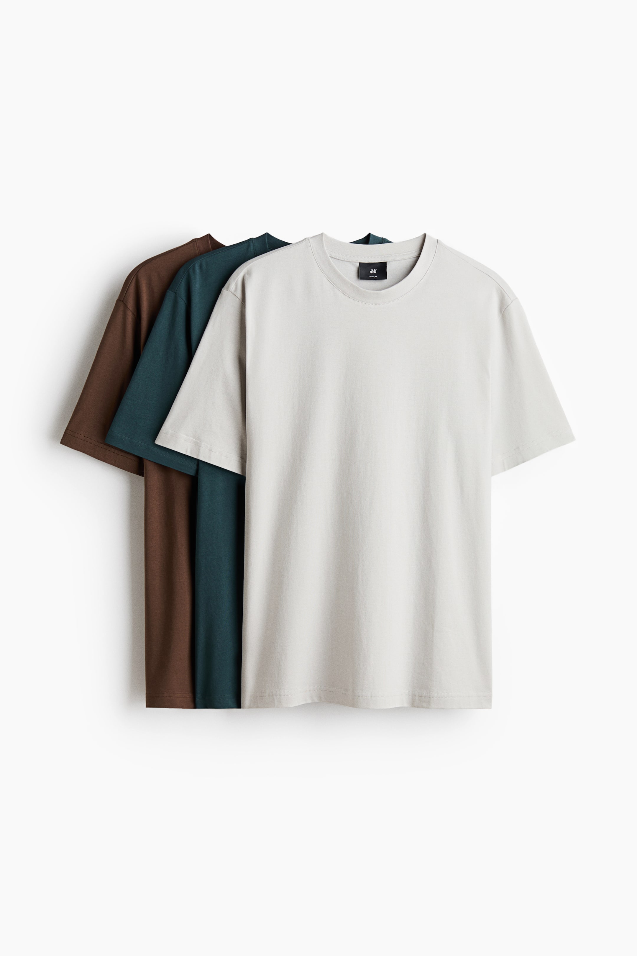 Agrandir l'image: Trois t-shirts sont présentés à l'AVANT, superposés avec un t-shirt gris clair à l'avant-plan, suivi d'un bleu canard foncé, puis d'un t-shirt brun foncé. Chaque t-shirt est confectionné en jersey de coton léger, avec un col rond bordé de côtes, un ourlet droit et une coupe régulière.