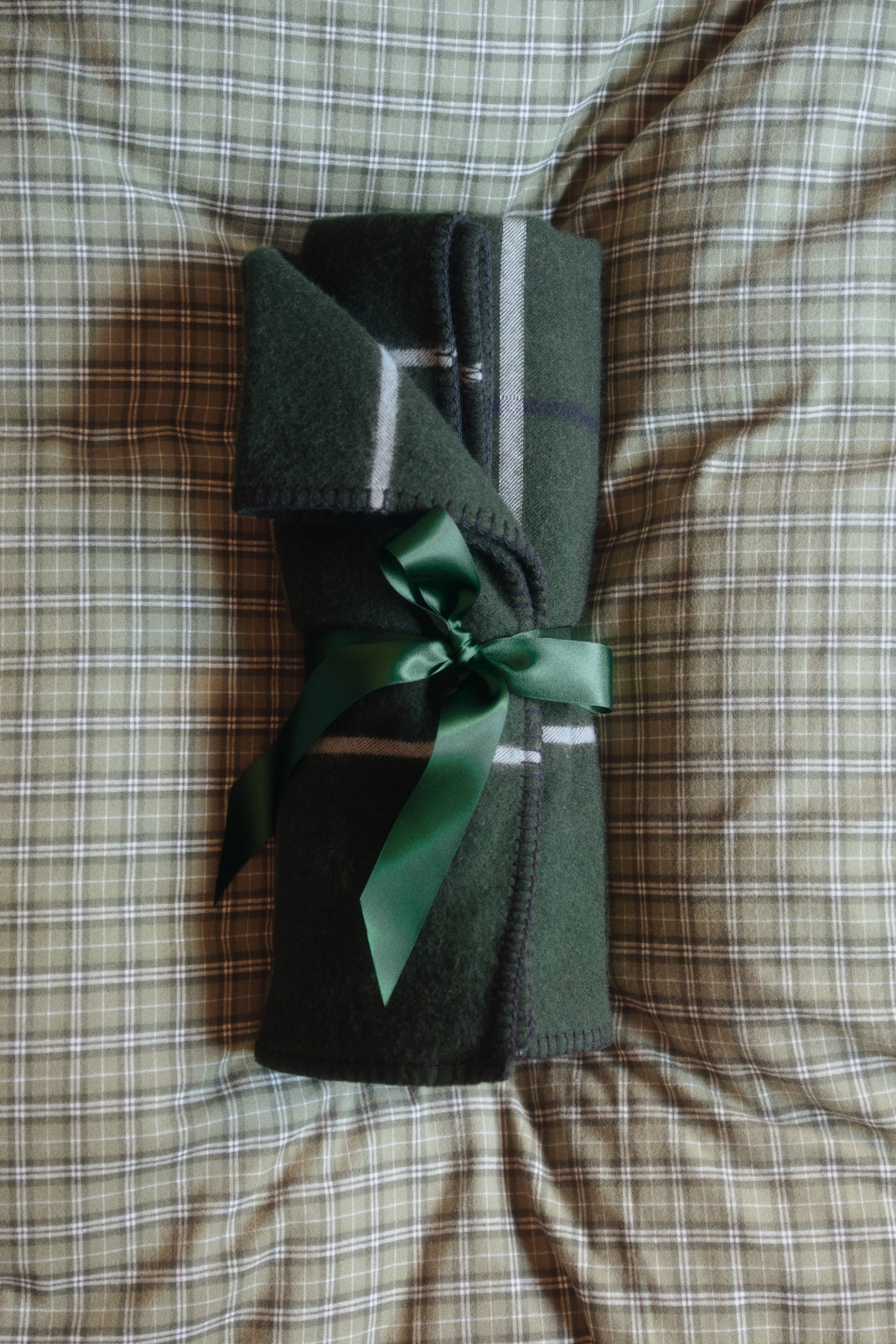 Agrandir l'image: Une couverture en mélange de laine vert foncé, roulée, présente un motif à carreaux blanc et des points de couverture sur ses bords. La couverture est maintenue au milieu par un ruban de satin vert assorti, noué en nœud.