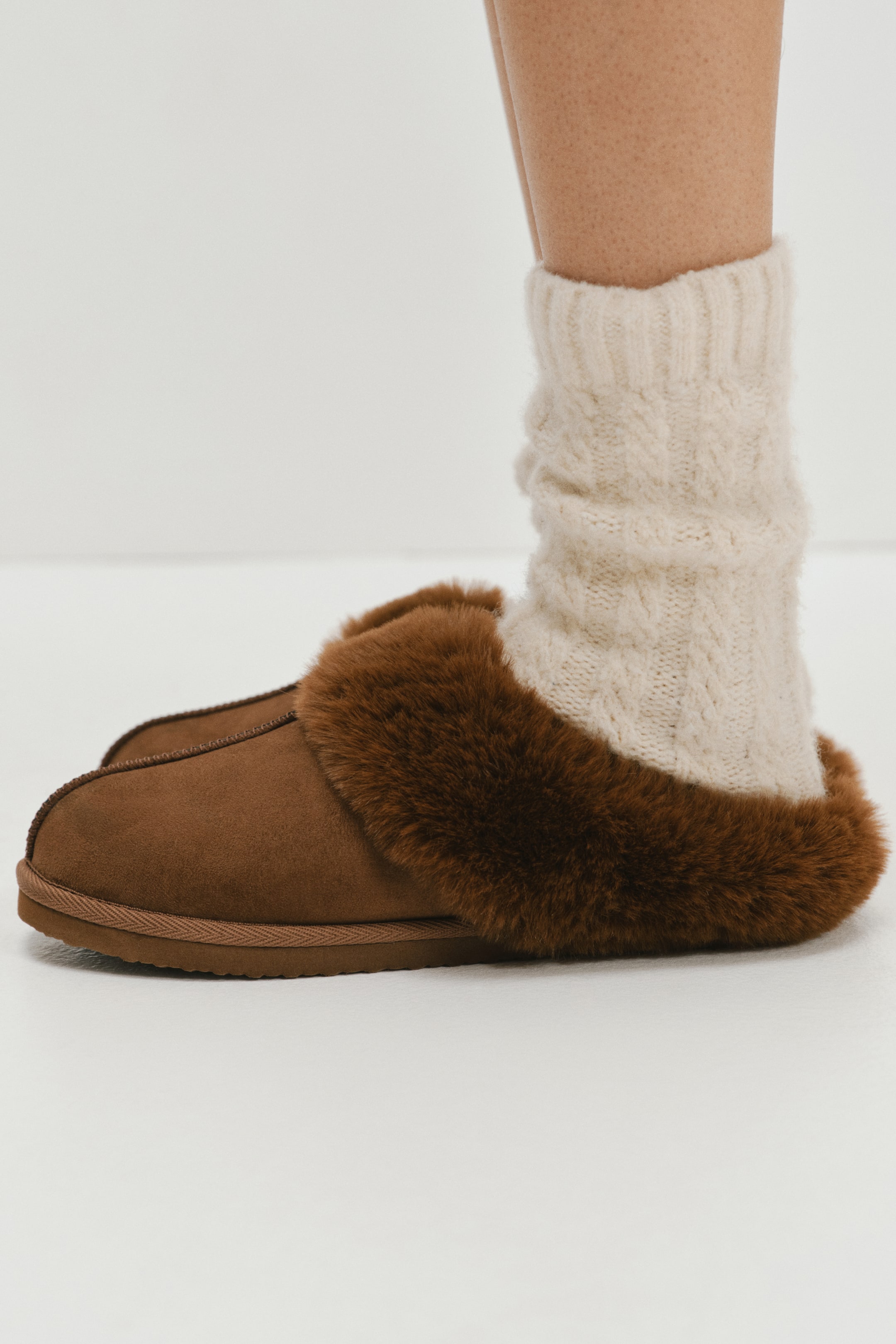 View larger image: Slippers with a brown napped finish and fluffy dark brown faux-fur lining are being worn by the model over cream-coloured cable-knit socks. The closed-toe, slip-on style features a flat textured sole and a plush collar around the foot opening.