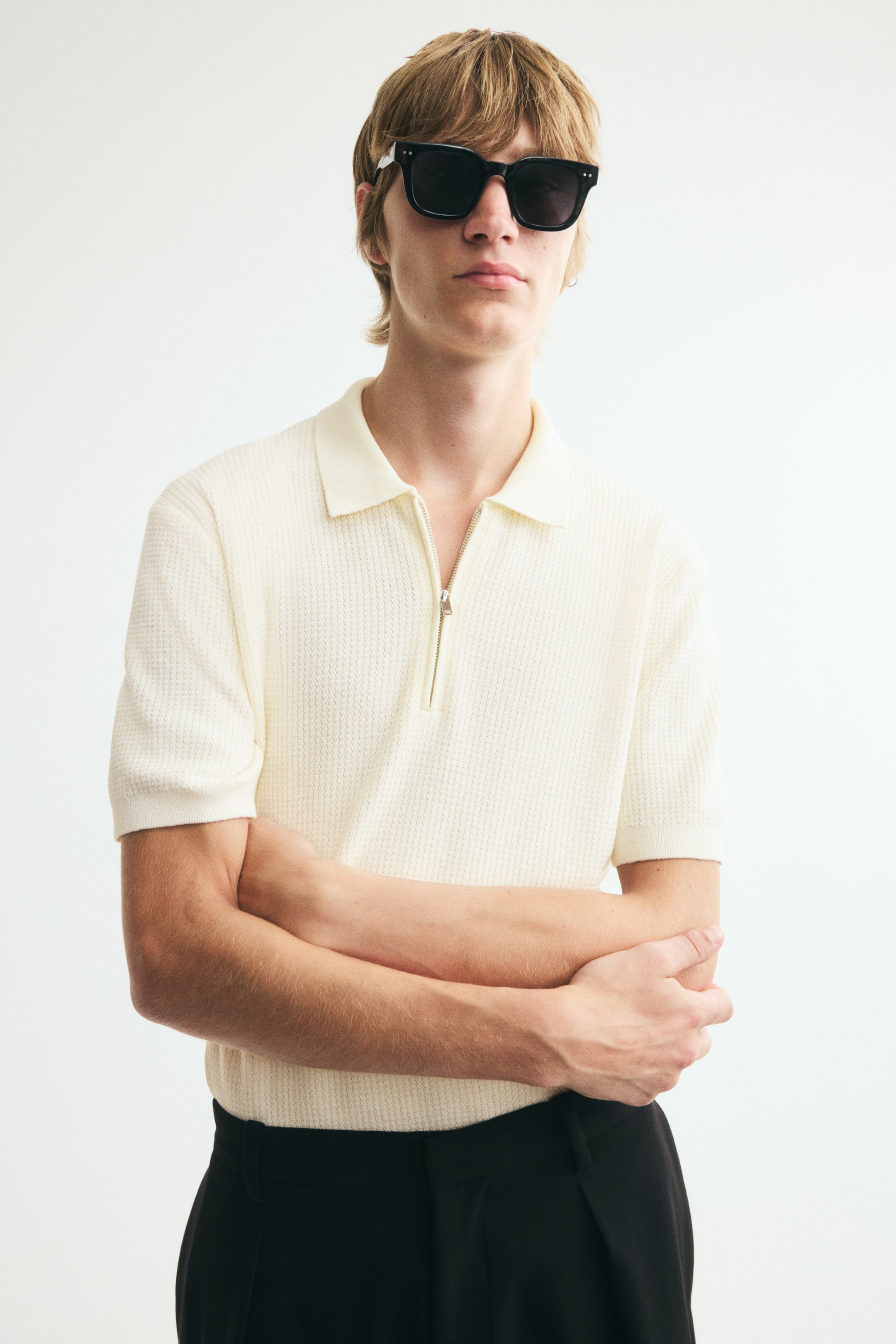 View larger image: A cream short-sleeved polo shirt in a textured knit is worn by the model, featuring a ribbed collar, a partially unzipped front, and fine ribbing at the cuffs and hem. The slim-fit shirt is tucked into black trousers, closely contouring the model’s body.
