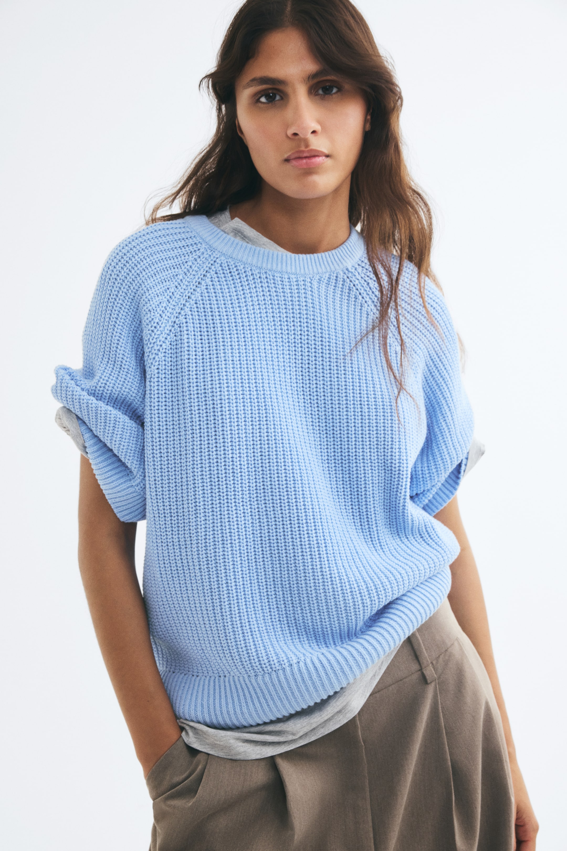 View larger image: An oversized light blue tee in a soft rib knit is worn by the model, layered over a grey top and paired with brown trousers. The tee features a round, rib-trimmed neckline, raglan sleeves rolled up at the shoulders, and a ribbed hem.