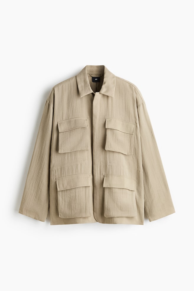 Eine beige Utility-Jacke ist von vorne zu sehen und prÃ¤sentiert einen strukturierten Stoff, einen klassischen Kragen und eine verdeckte Frontleiste. Sie verfÃ¼gt Ã¼ber vier groÃe, symmetrisch angeordnete Pattentaschen auf der Vorderseite sowie lange, lockere Ãrmel. - 1