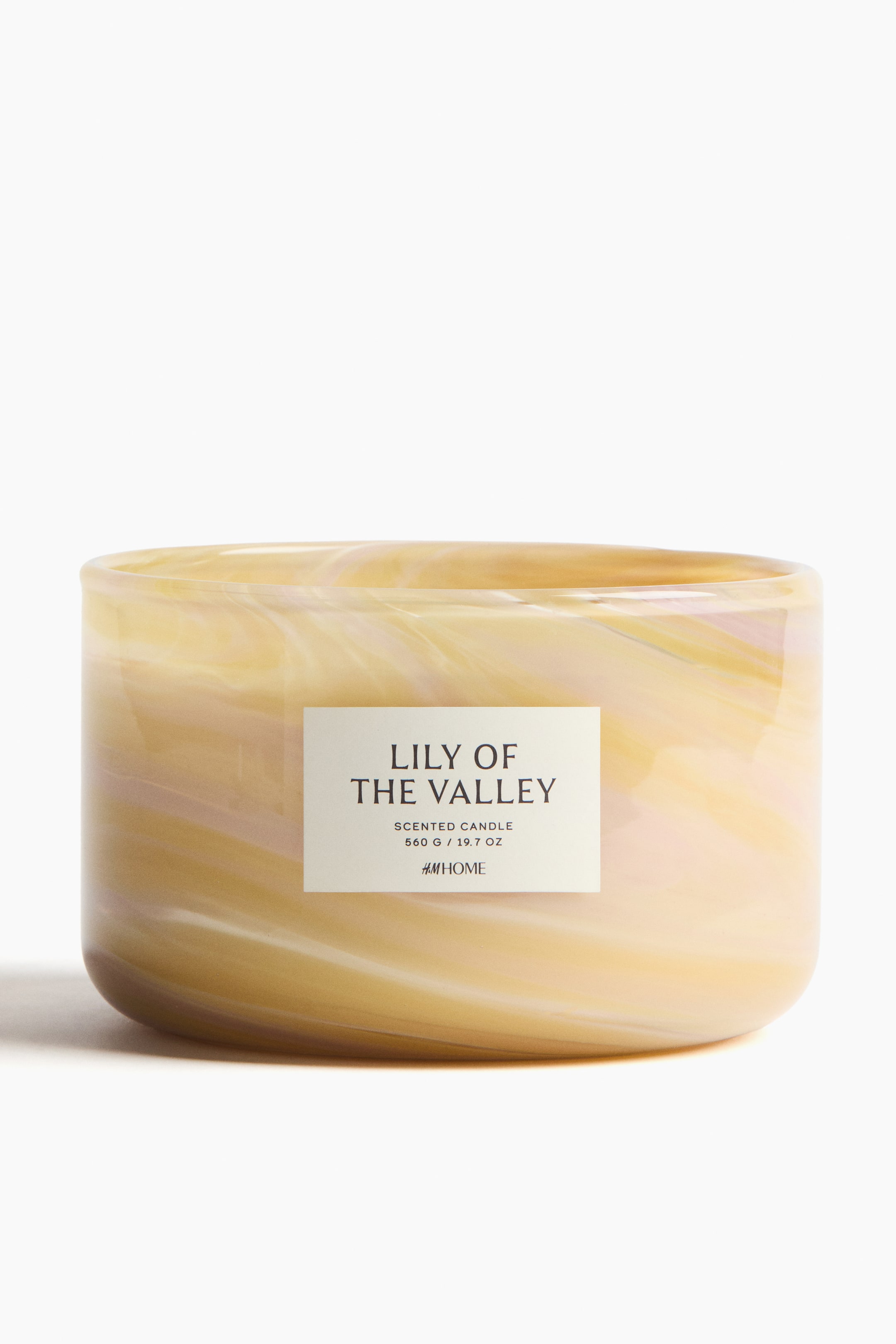 View larger image: A front-facing scented candle is housed in a glass holder, featuring a swirled, marbled pattern in pale yellow and cream tones. A rectangular label on the front displays "LILY OF THE VALLEY", "SCENTED CANDLE", "560 G / 19.7 OZ", and "H&M HOME".