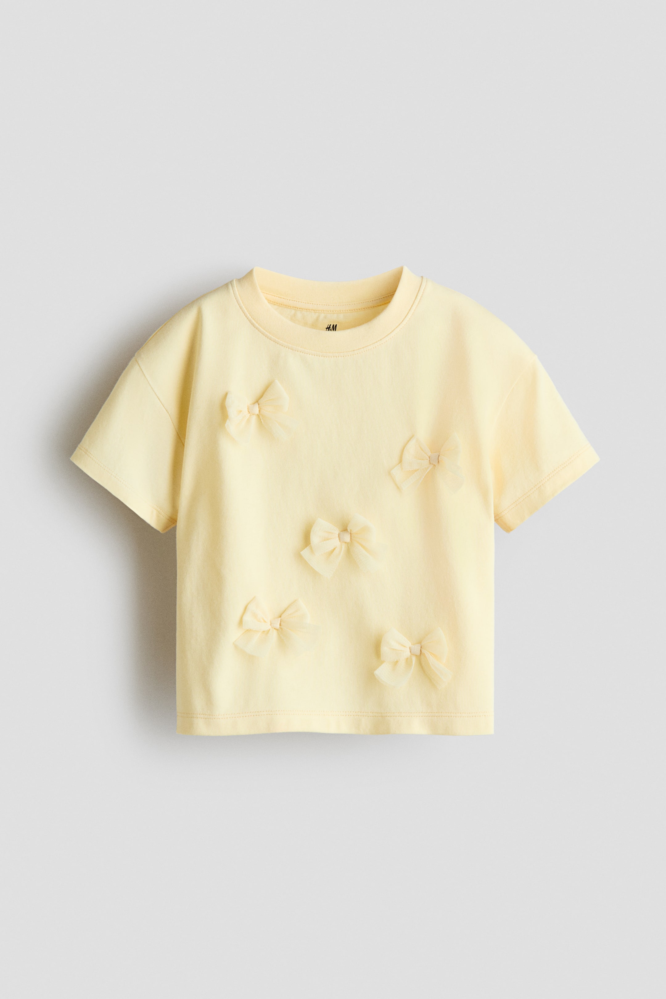 Größeres Bild anzeigen: Ein hellgelbes, locker sitzendes T-Shirt wird von vorne präsentiert, mit einem gerippten Besatz am Halsausschnitt und tief angesetzten Schultern. Mehrere kleine, transparente Schleifen zieren die glatte Baumwolljersey-Vorderseite.