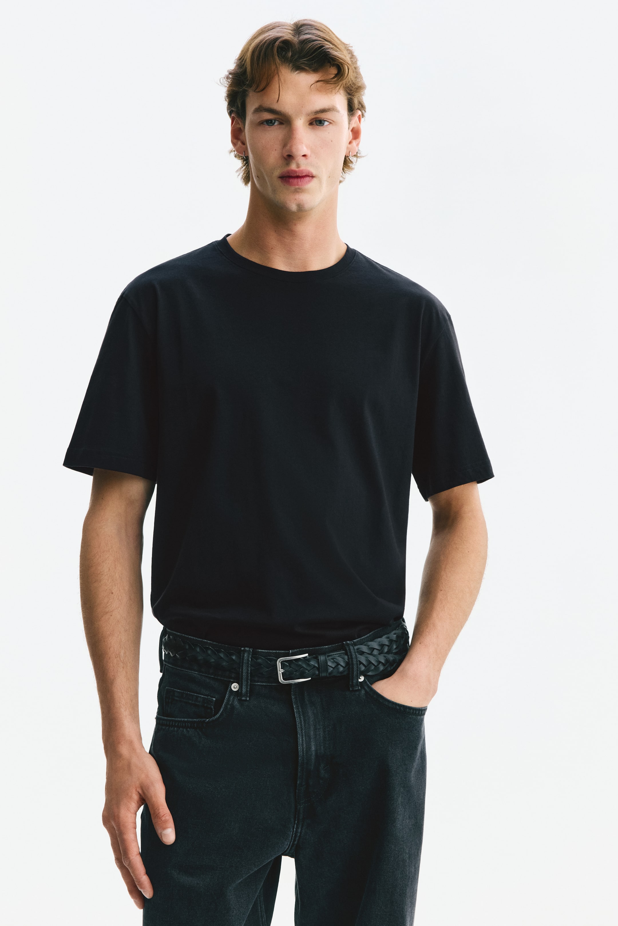 View larger image: Model tersebut memakai T-shirt hitam dengan leher bulat, lengan pendek, potongan biasa, dan hem lurus. Model berdiri dengan satu tangan di dalam poket seluar gelap dan memakai tali pinggang anyaman.
