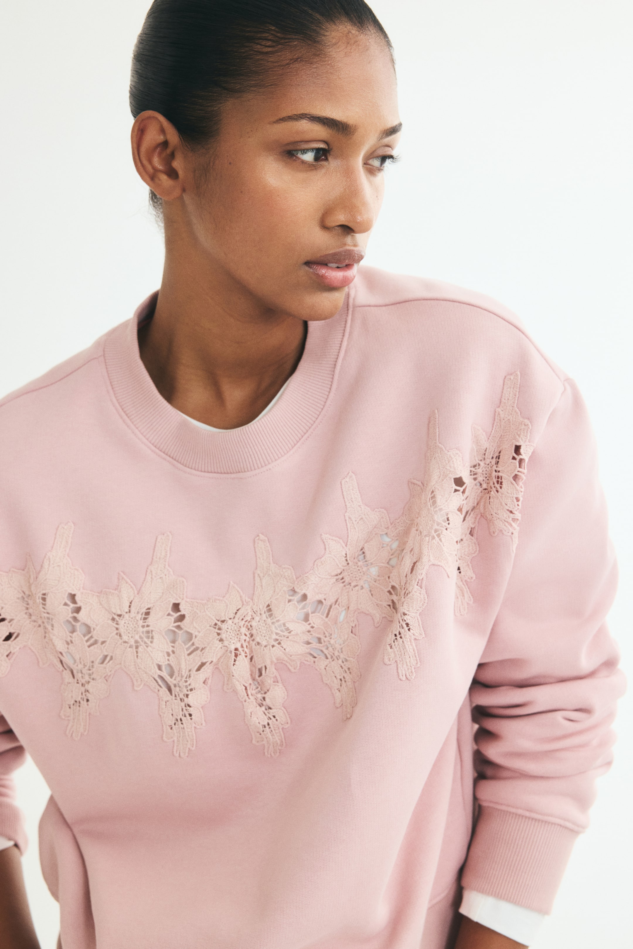 View larger image: A pink, loose-fit sweater is shown from the front, featuring an irregular floral lace inset detailed across the chest. The sweater is designed with a ribbed round neckline, dropped shoulders, and ribbed cuffs, with a white underlayer peeking out at the neck and wrist.
