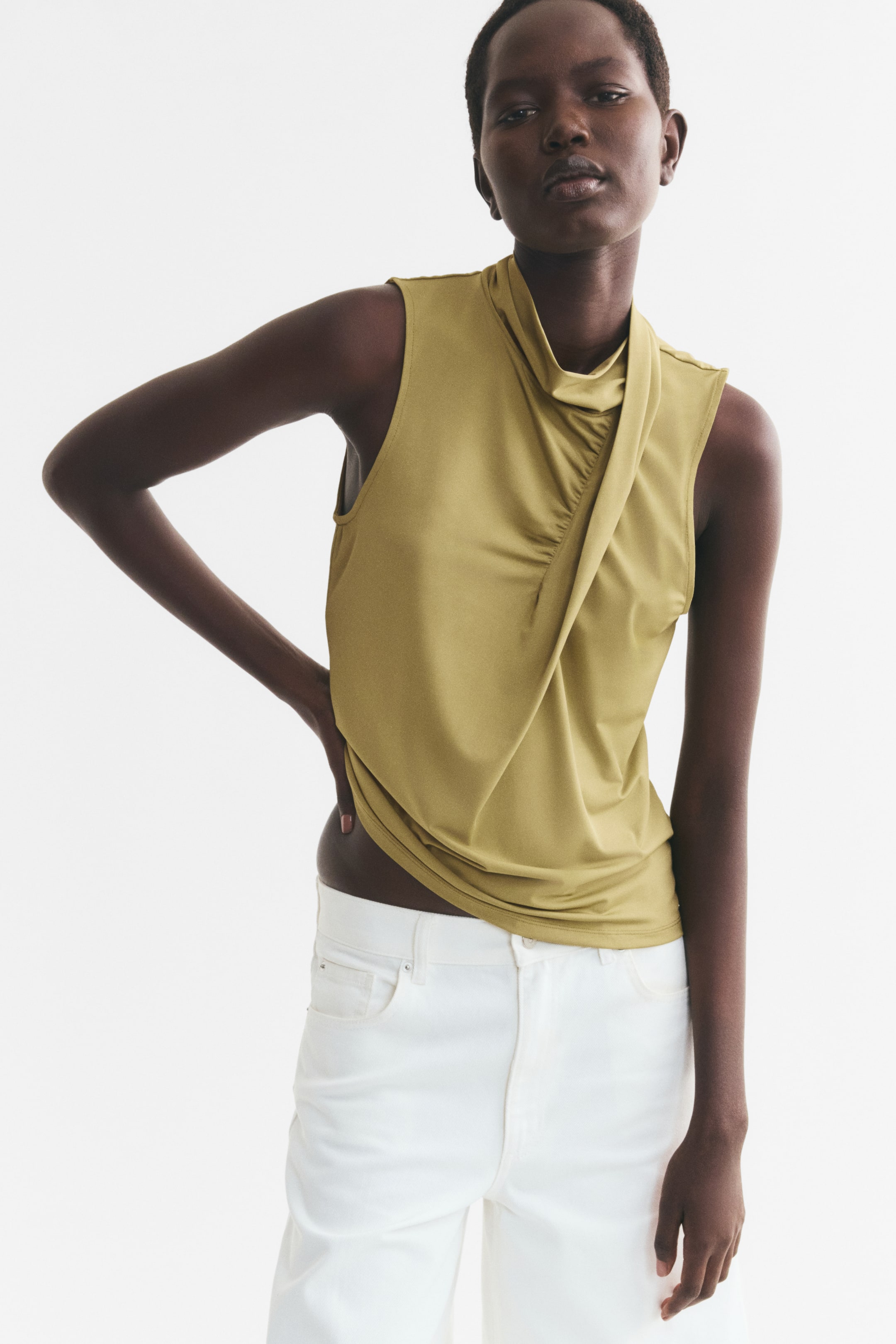 View larger image: A sleeveless, olive-green stretch jersey top is being worn by the model, featuring a double-layered stand collar that transitions into a sewn-in, draped panel down the front. The model is standing with one hand resting on their hip, styling the top with white trousers.