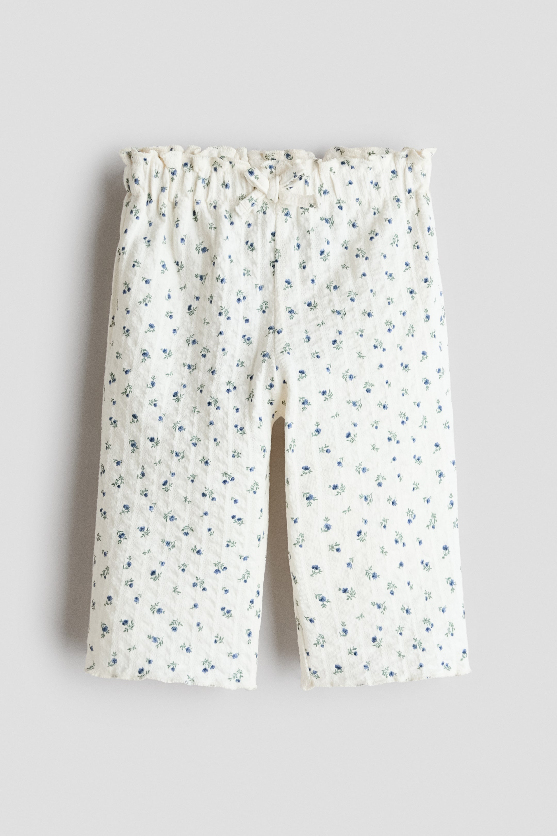 Agrandir l'image: Le pantalon est présenté de face, mettant en avant un tissu en jersey crème texturé avec un petit imprimé floral bleu. Il est conçu avec une taille ornée de volants, affichant un petit nœud à l'avant, et tombe avec une coupe ample au niveau des hanches et des cuisses, se terminant par des jambes larges avec des ourlets surjetés.