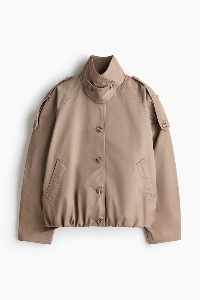 Um casaco bomber em cor taupe exibe uma gola alta com abas abotoadas, mangas raglan com dragonas abotoadas e um fecho frontal completo com botões. Bolsos de debrum angulados são visíveis na frente, e a bainha é elástica. - 1