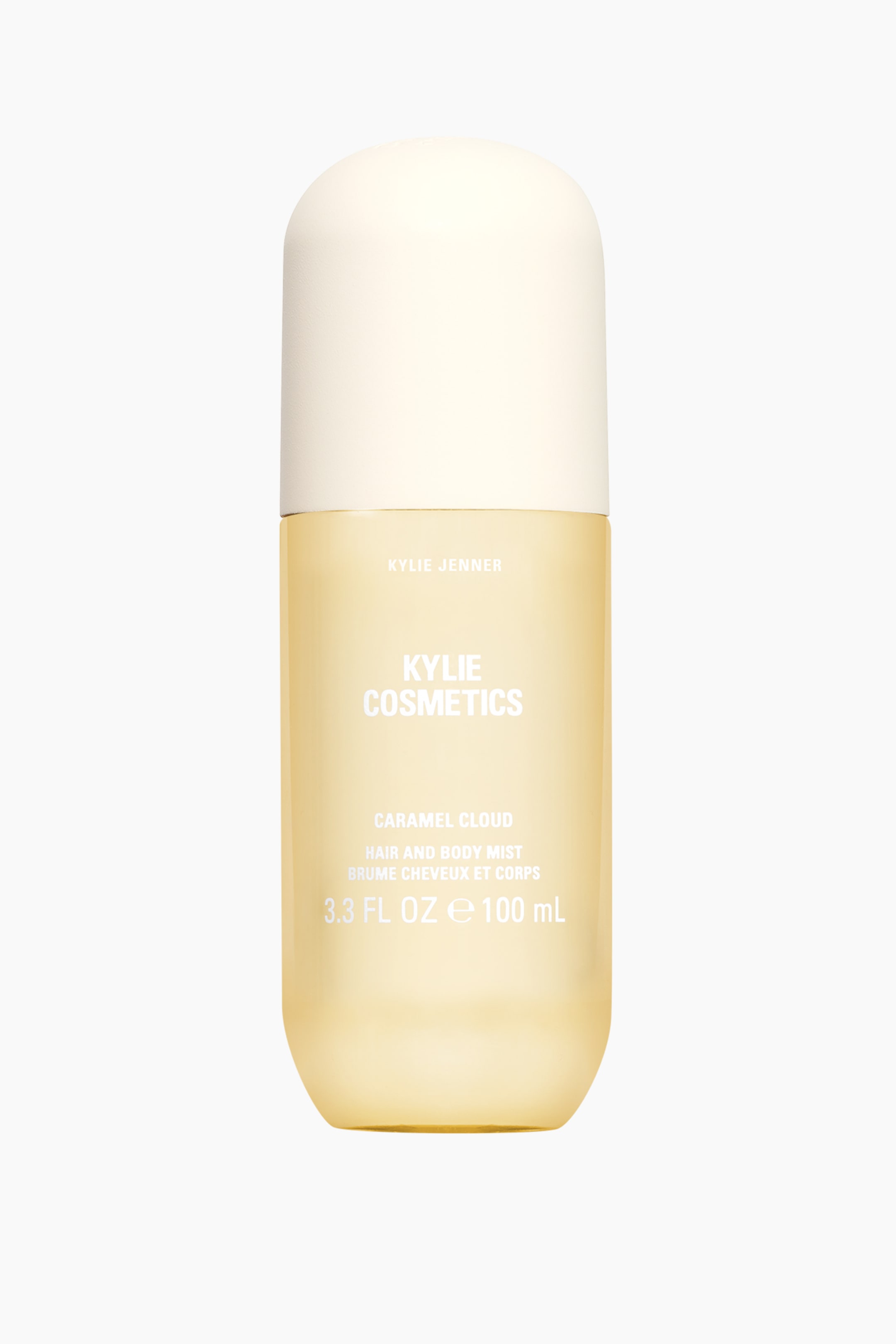 Se større billede: En cylindrisk, gennemsigtig ravfarvet flaske med Kylie Cosmetics Caramel Cloud Hair and Body Mist står oprejst og har en cremefarvet kuppelformet hætte. Hvid tekst er trykt lodret på flasken og angiver "Kylie Jenner," "Kylie Cosmetics," "Caramel Cloud," "Hair and Body Mist" og "3.3 fl oz e 100 mL."