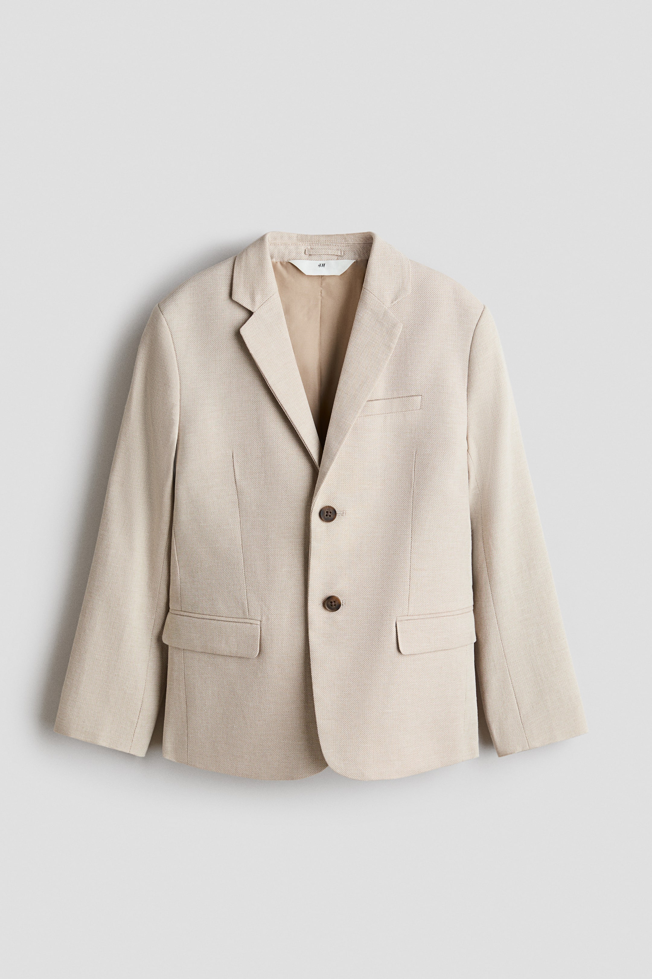 View larger image: A beige blazer is shown from the front, featuring a lightly textured weave, notch lapels, a two-button closure with brown buttons, one chest welt pocket, and two flap pockets.