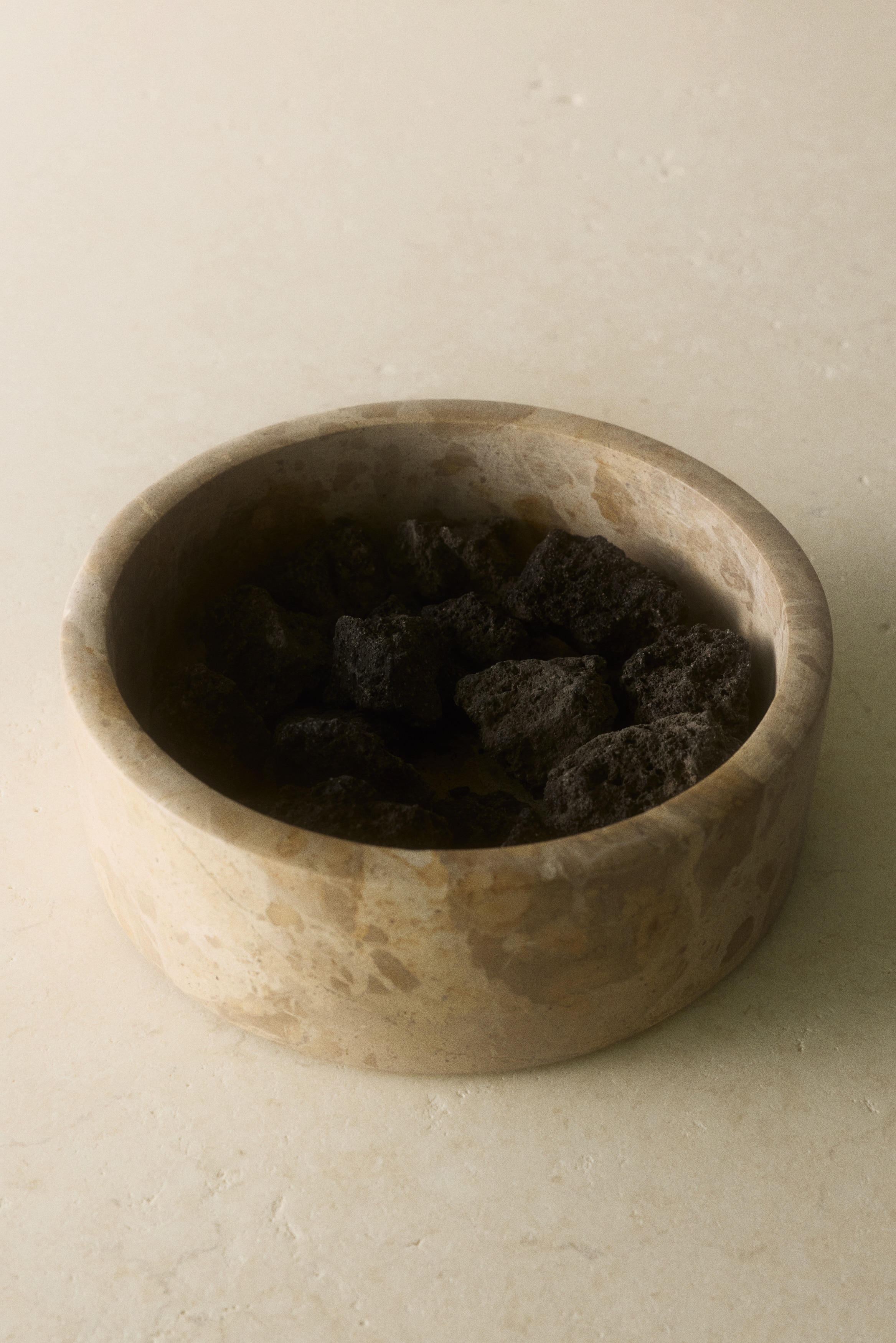 A round beige marble container with a flat matching lid is positioned facing front, showing natural brown and tan veining. Three small textured black lava rocks are resting on the surface directly to its right.