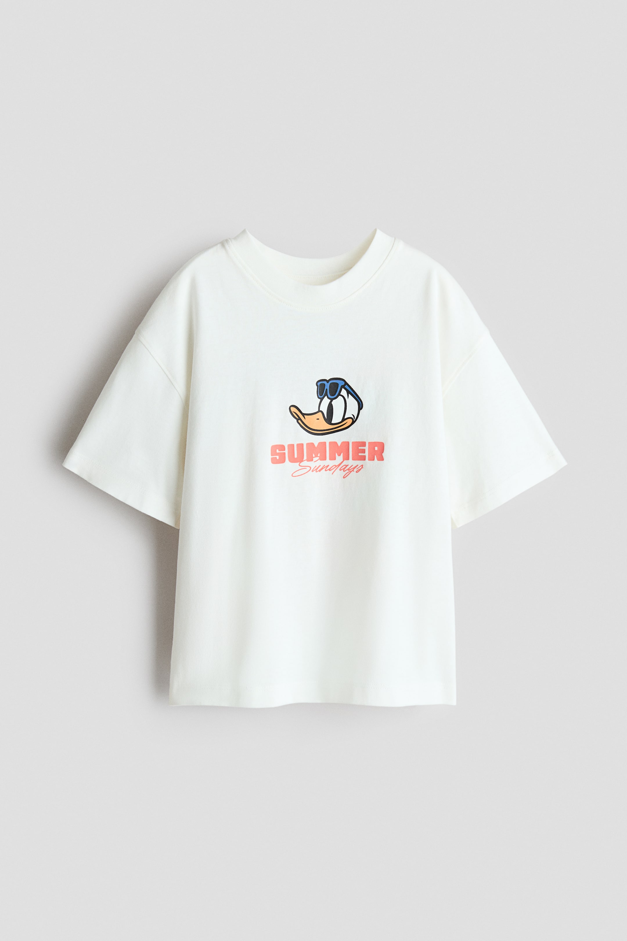 View larger image: A cream-coloured, oversized t-shirt crafted from soft cotton jersey is displayed front-facing. It features ribbed trim around the neckline, dropped shoulders, and a graphic print on the chest depicting a duck wearing blue sunglasses above the words 'SUMMER' in coral and 'Sundays' in a darker coral cursive.