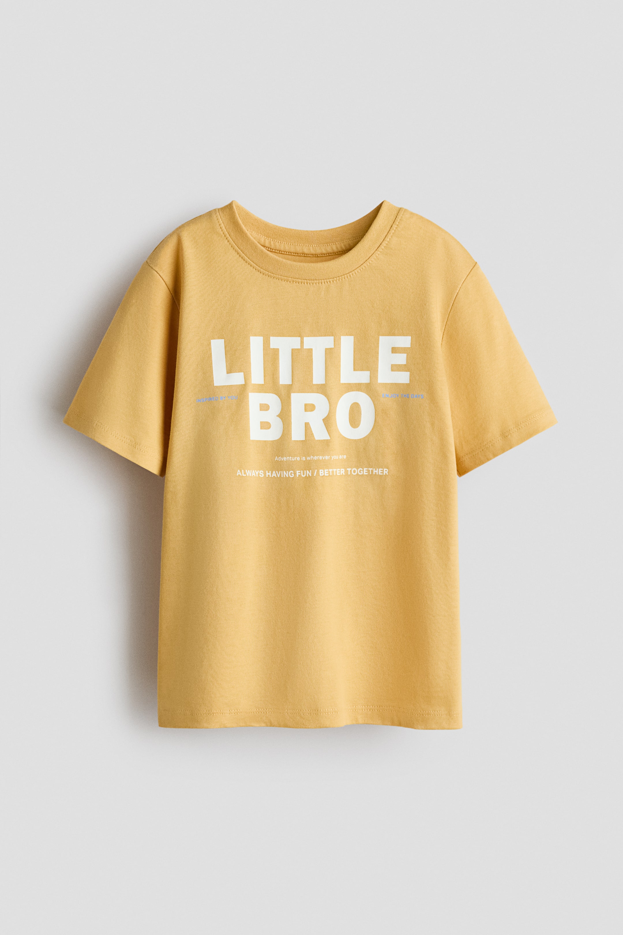 View larger image: A front-facing T-shirt, made from soft cotton-jersey, in a mustard-yellow colour with ribbing around the neckline, dropped shoulders, and a straight-cut hem. The front bears the white text "LITTLE BRO" in large letters, accented by smaller white phrases above and below.