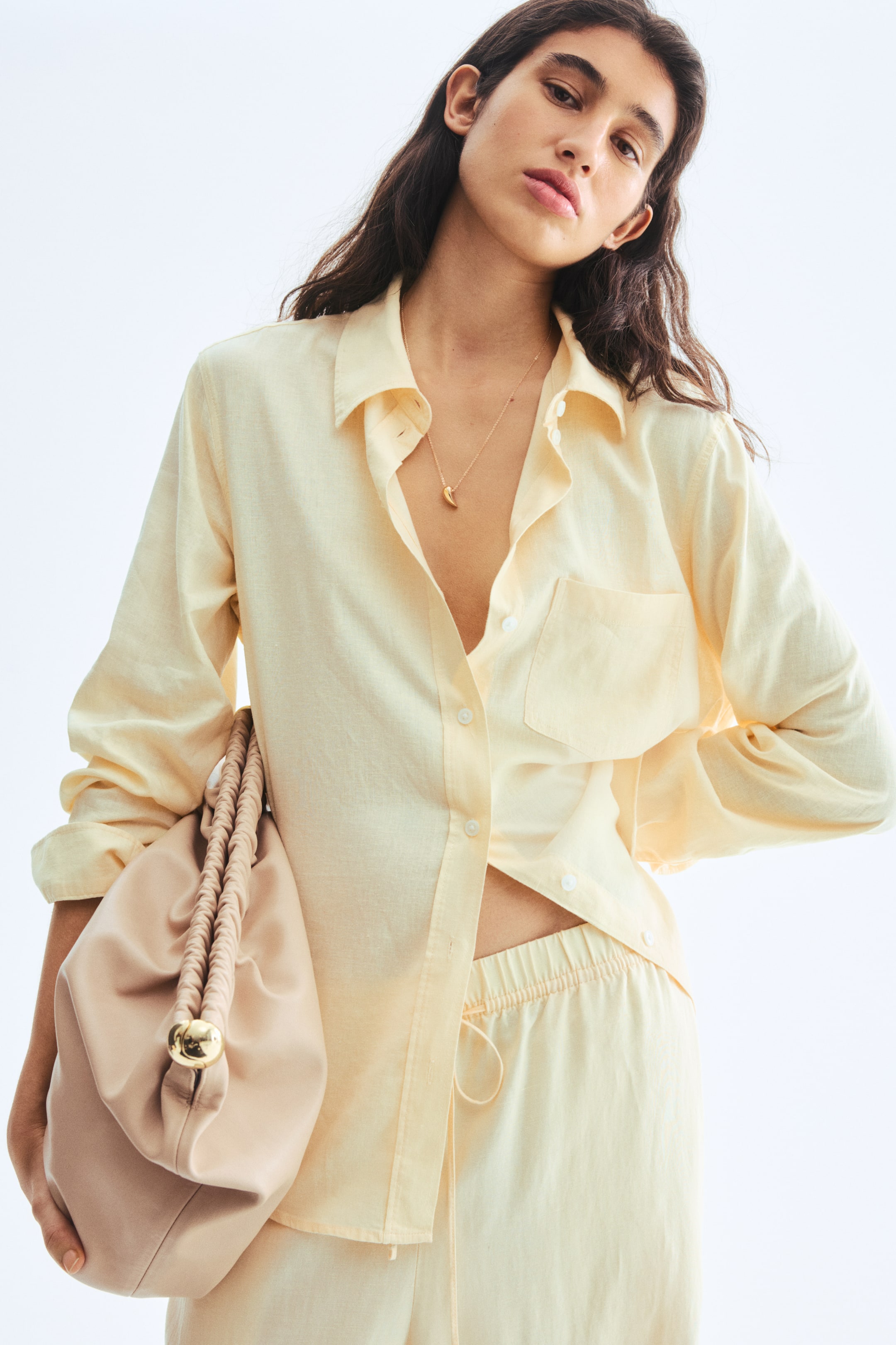 View larger image: A pale yellow long-sleeved button-up shirt, featuring a pointed collar and a chest patch pocket, and matching relaxed-fit pants with an elasticated drawstring waist are being presented from a slightly low angle, with the shirt styled unbuttoned at the neck and untucked.
