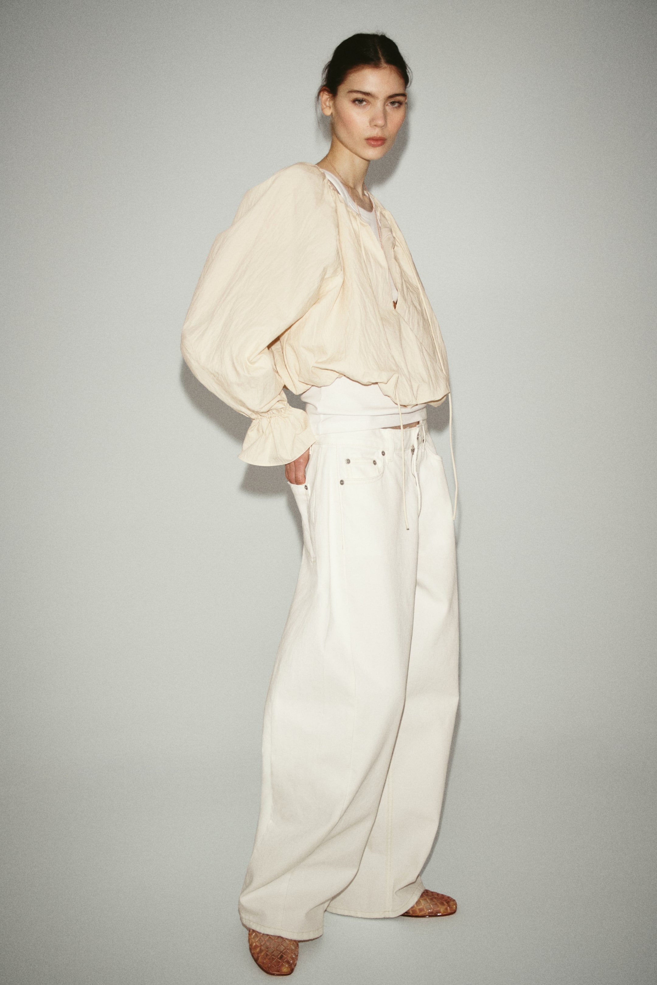 View larger image: A light beige crinkled cropped jacket features voluminous long sleeves with ruffled cuffs and a drawstring hem, layered over a white inner top. Light cream wide-leg pants complement the jacket, showcasing visible rivets and a drawstring waist, while the entire ensemble displays a relaxed silhouette from a slight angle, with brown textured slip-on shoes completing the look.
