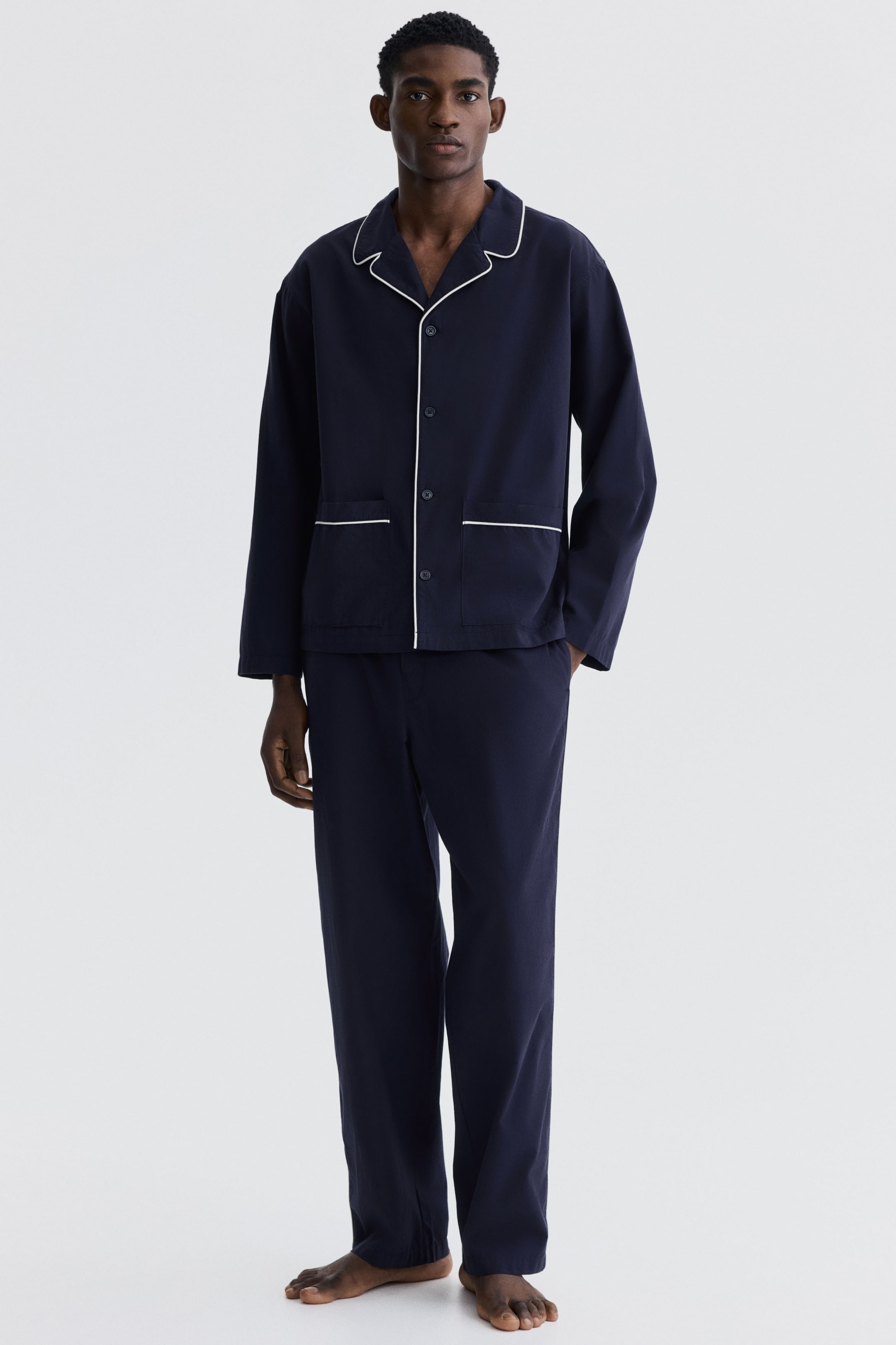 Ampliar la imagen: Se presenta un conjunto de pijama para hombre en color azul marino, que incluye una camisa de manga larga con solapas de muesca, ribetes blancos a lo largo de las solapas, la tapeta delantera y dos bolsillos de parche, acompañado de pantalones largos a juego.