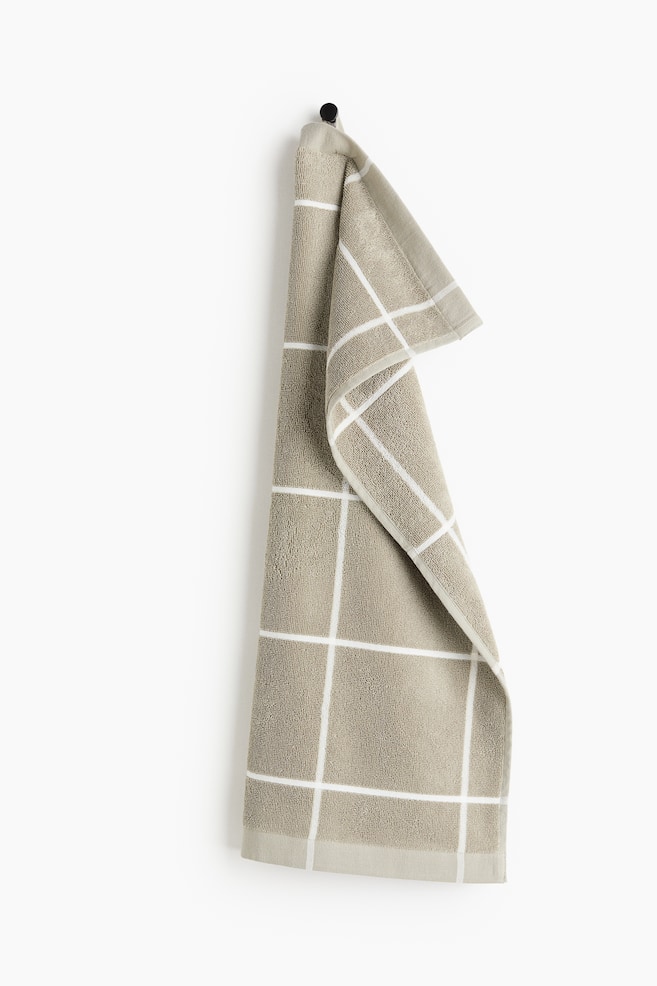 Une serviette en tissu éponge beige avec un motif à carreaux blancs est suspendue à un crochet noir, pliée et retombant vers le bas. - 1